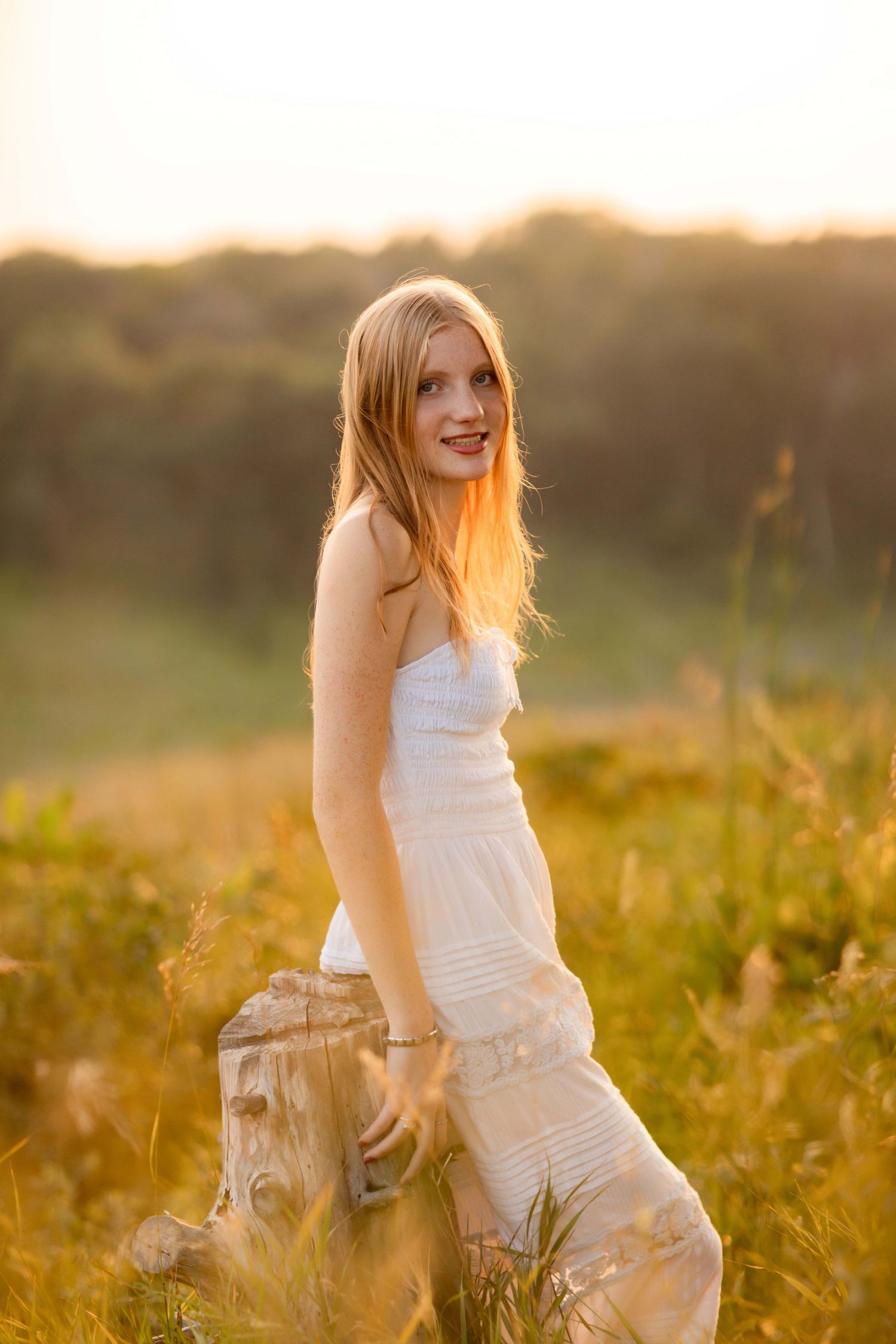 Young woman with long blonde hair in white sundress posing outdoors in golden sunset light in a meadow.