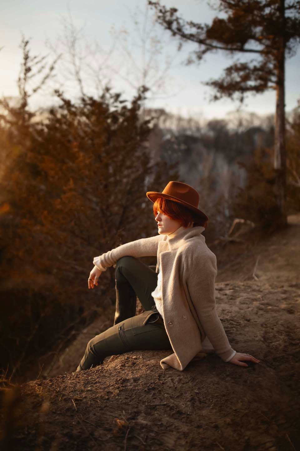 A person in winter attire sits contemplatively on a cliff edge during golden hour.