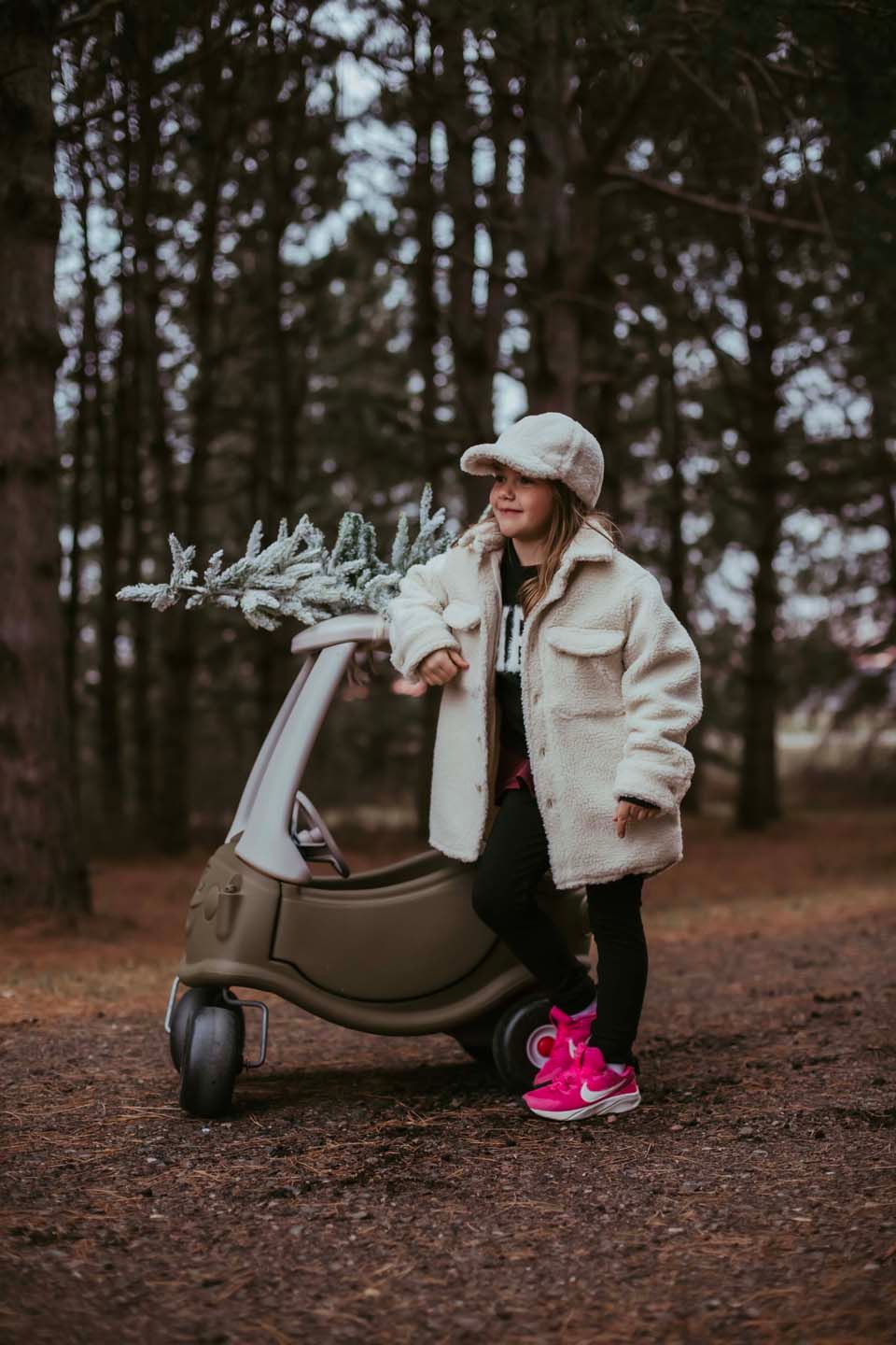 Child in cozy winter clothing playing with toy car and festive decorations in forest.