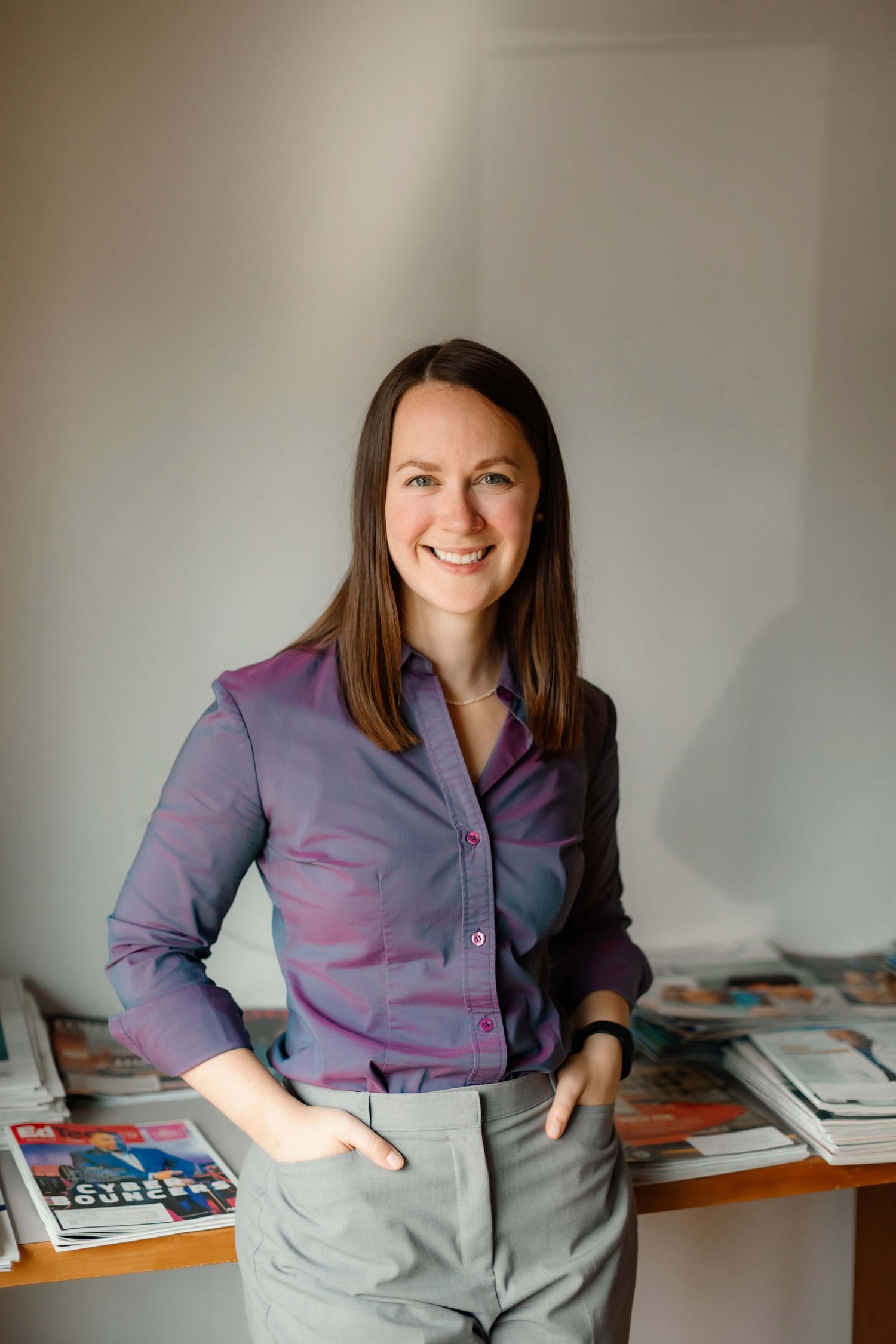 Professional wearing purple button-down shirt and gray pants stands confidently next to desk with books.