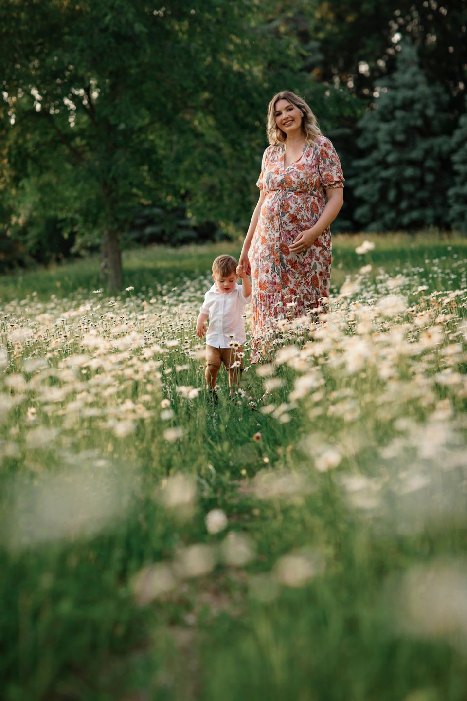 A mother and child stand together in a dreamy summer field of white wildflowers during golden hour.
