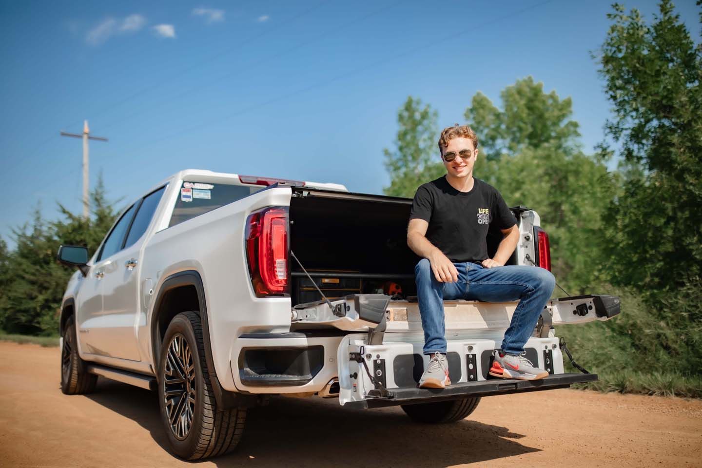 Person sitting casually in back of silver GMC pickup truck on dirt road with trees in background.