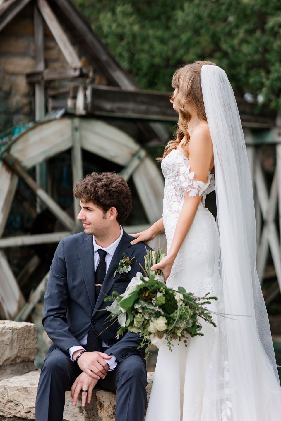 A romantic wedding portrait showcasing a bride in white lace and groom in navy against a rustic wooden backdrop.