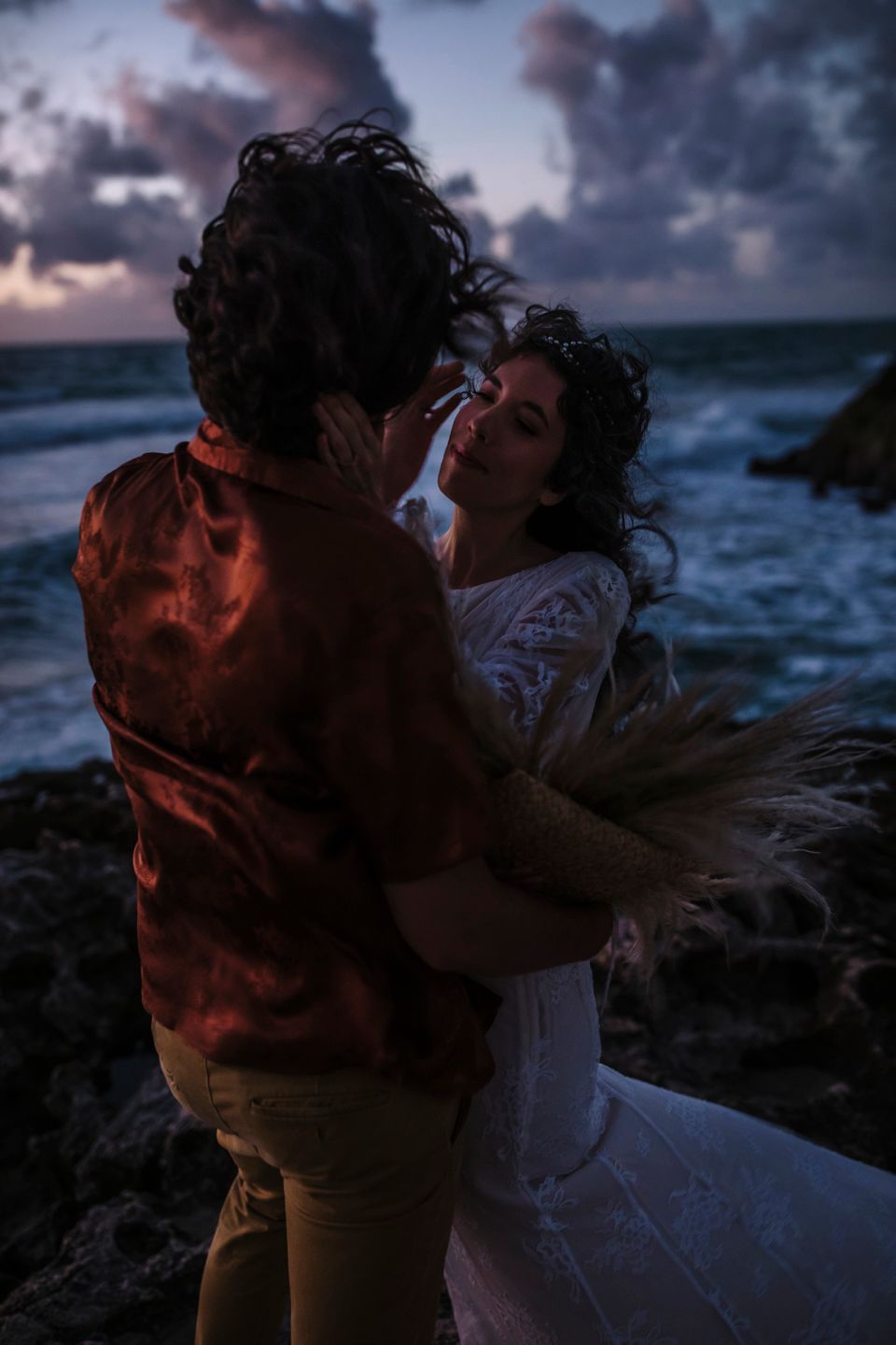 A romantic couple embraces on rocky coastal cliffs at sunset with dramatic clouds and crashing waves in the background.