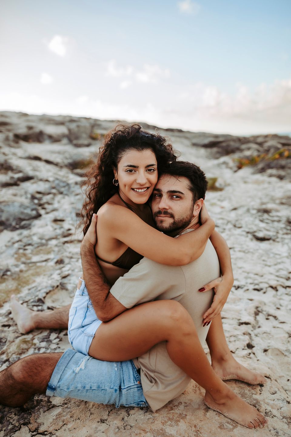 A couple embracing on rocky coastal terrain with ocean waves in the background.