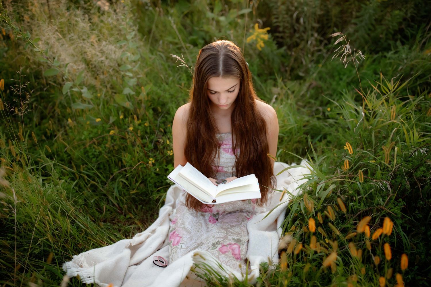 Reading a book in a flowing white floral dress amidst tall grass and wildflowers.