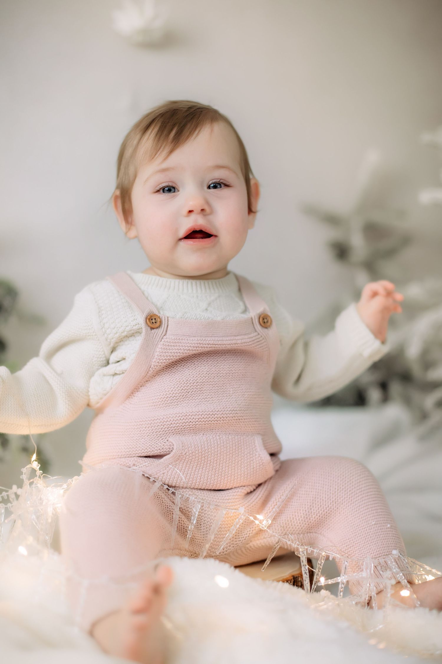 Baby in pink overalls and white sweater sits among twinkling lights with winter backdrop.