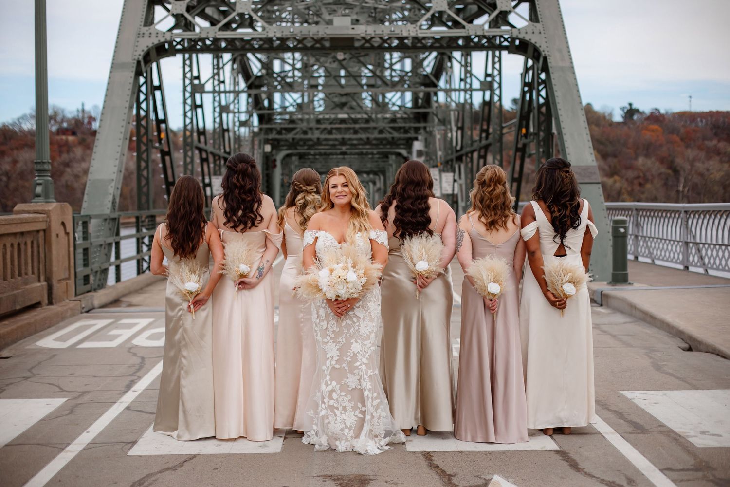 Wedding party in neutral-toned dresses standing on bridge with industrial architecture.