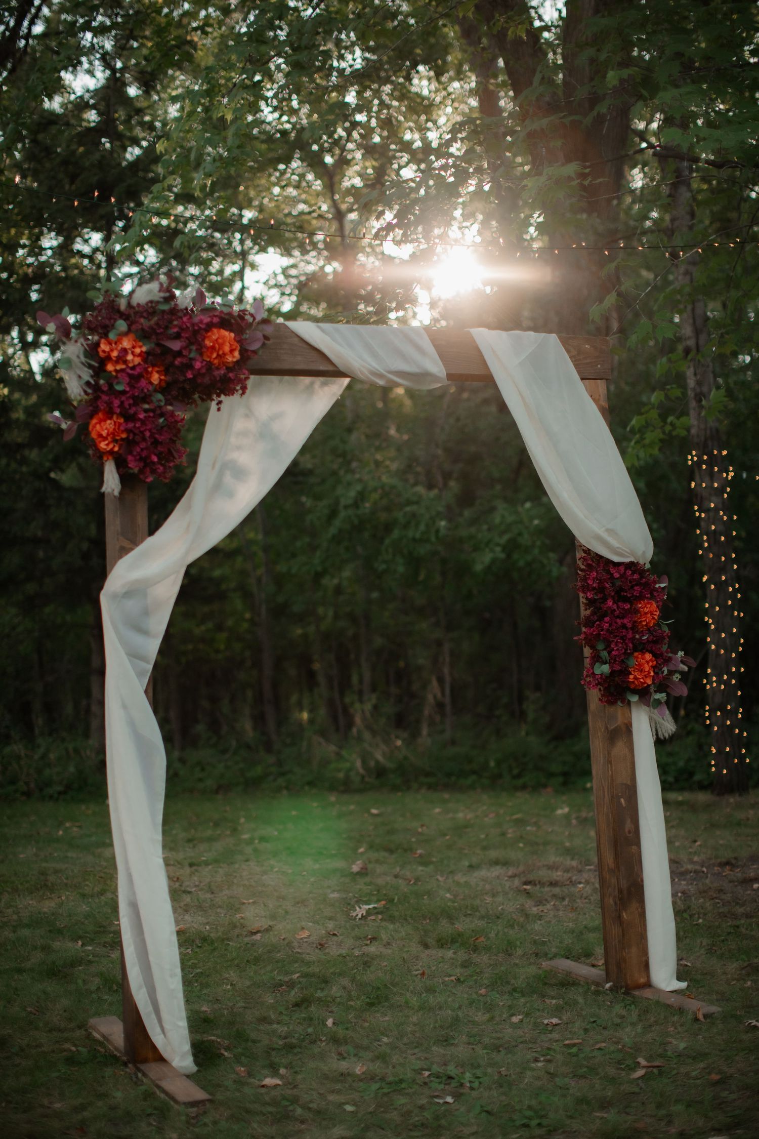Wedding arch decorated with red flowers and white fabric in a wooded setting with sunlight streaming through.