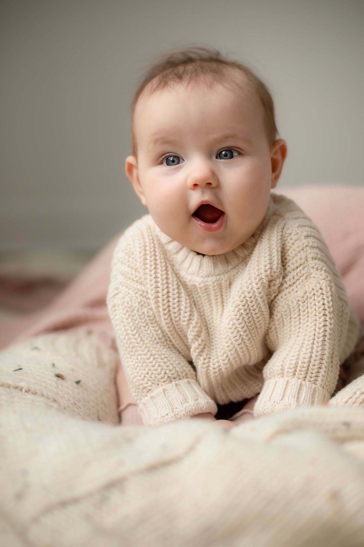 A baby wearing a cream knit sweater lies on soft pink bedding while making various playful expressions.