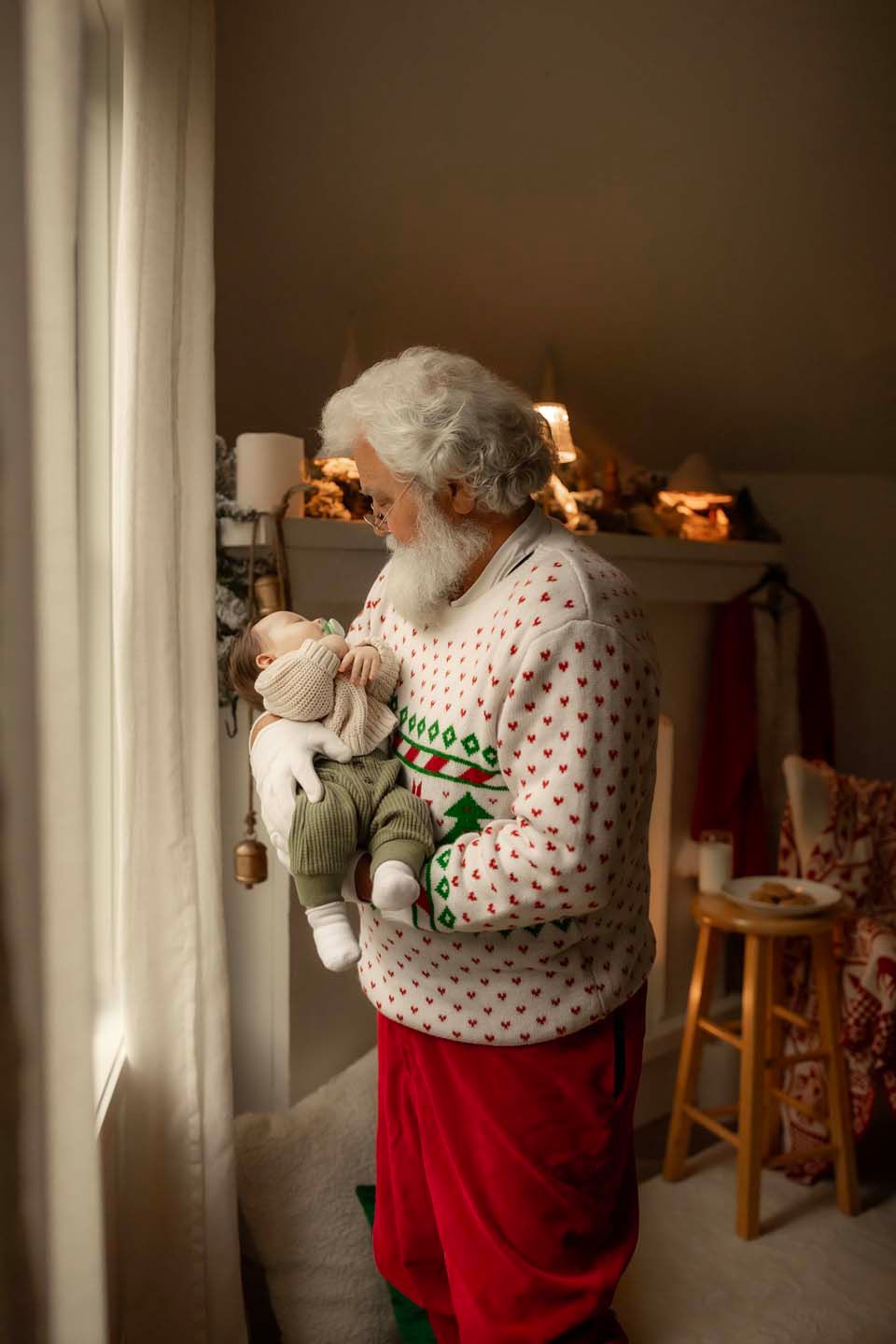 Santa Claus in festive sweater and red pants gently holds a newborn baby near a window with Christmas decorations.