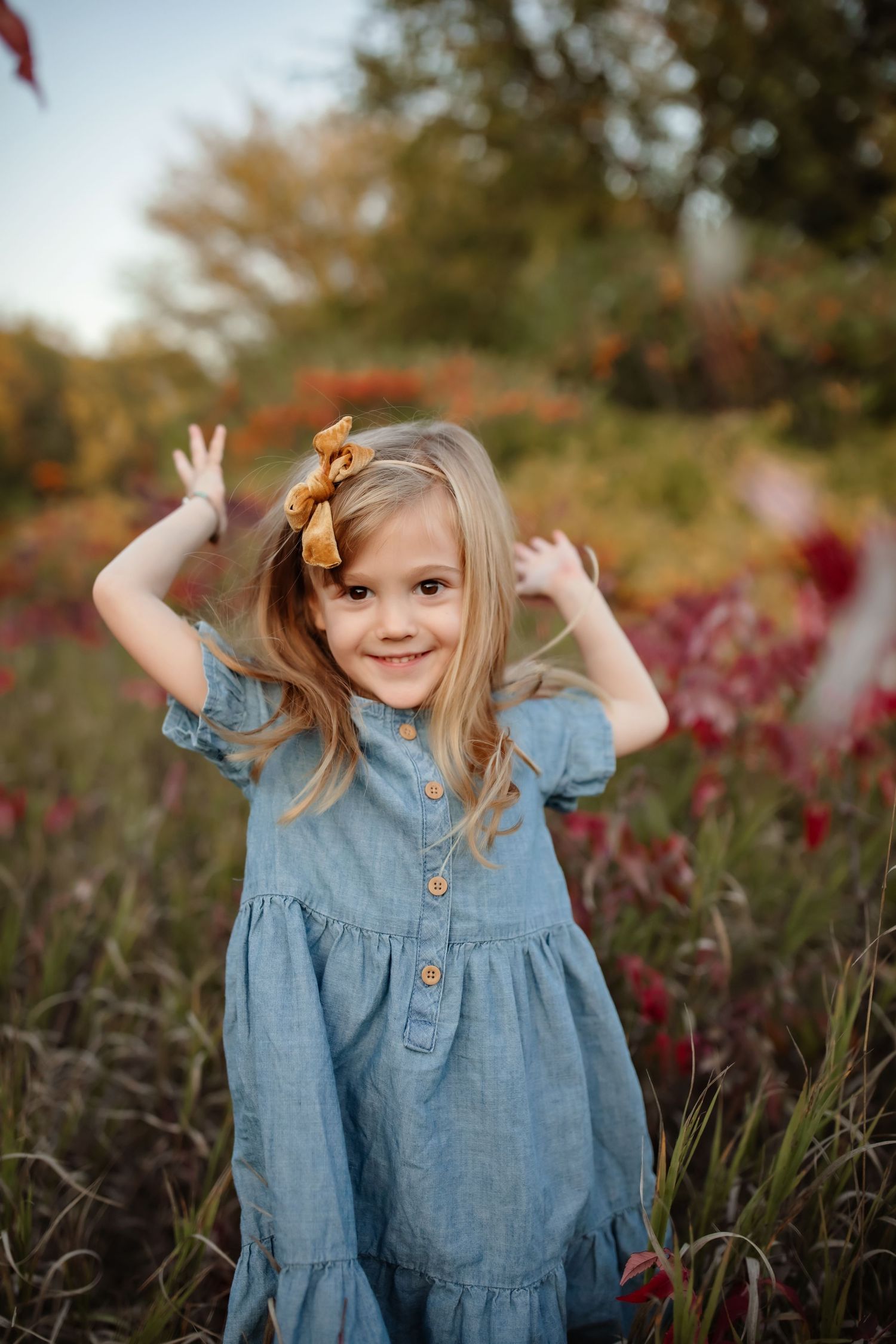 A child in a blue denim dress and golden bow plays in a garden with autumn foliage in the background.