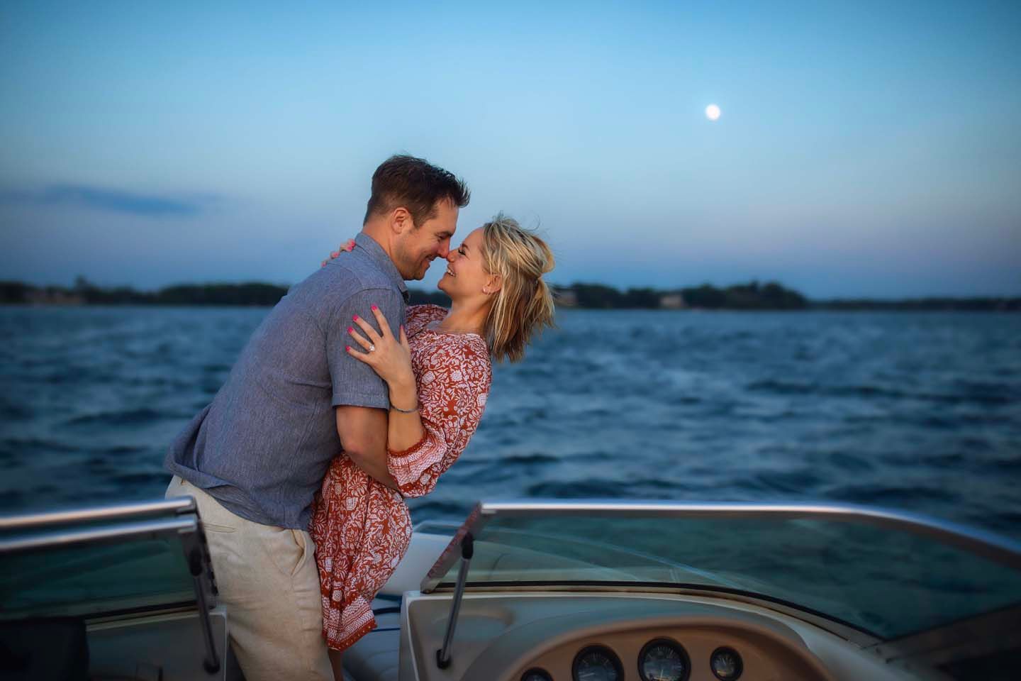 A romantic moment between a couple kissing on a boat at dusk with a moon visible in the blue sky.