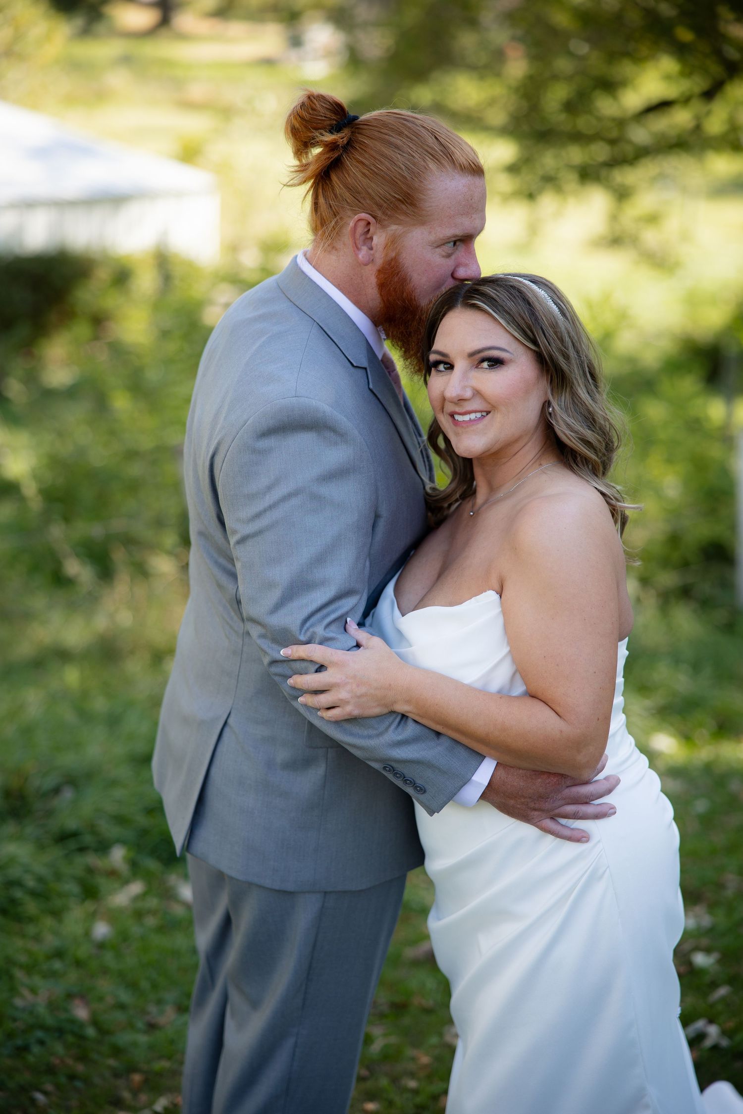 A couple shares a tender moment in an outdoor setting with sunlit trees in the background.