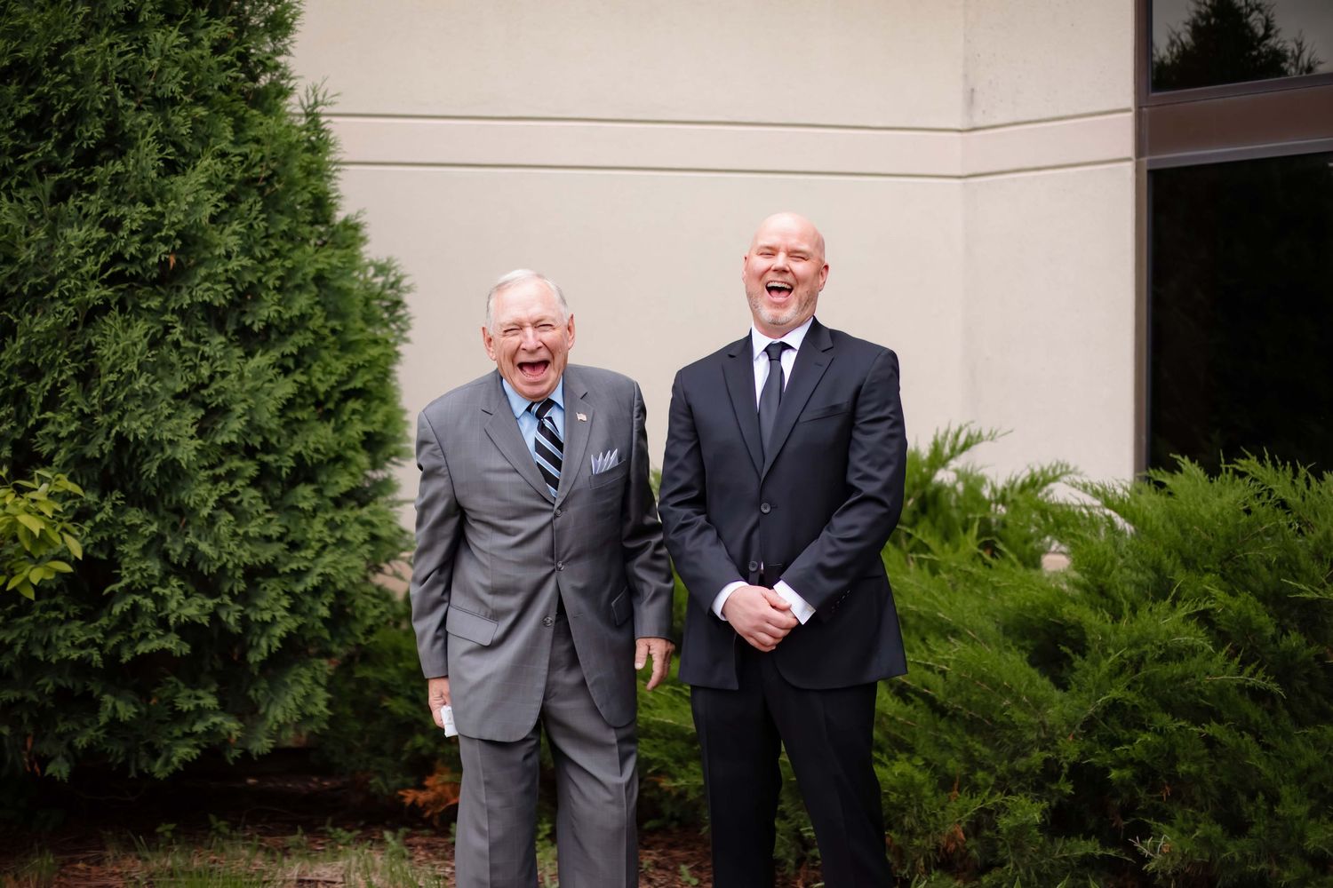 Two people in business suits stand together smiling outside a building with greenery in the background.