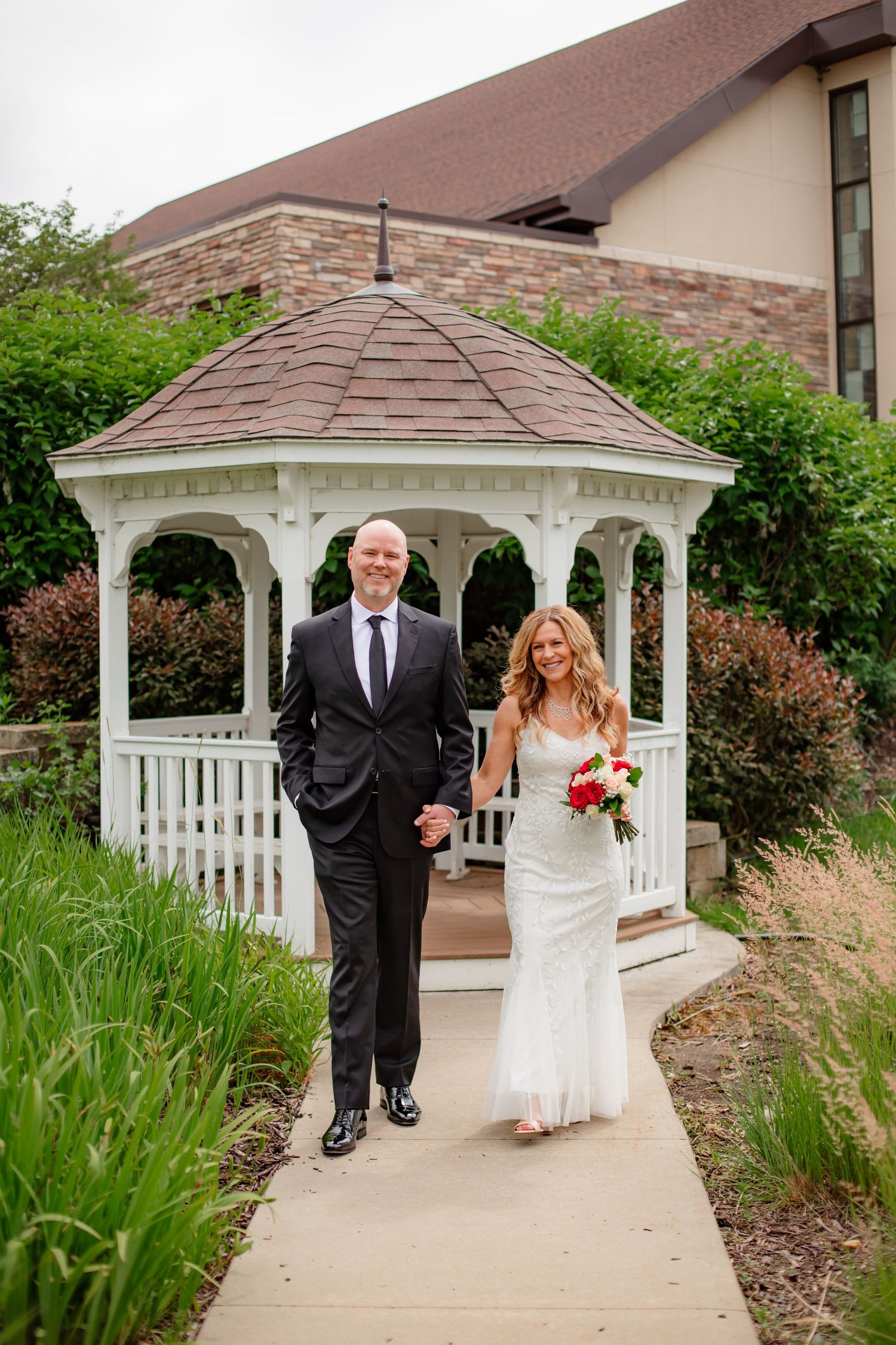 A wedding couple poses for photos in front of a white garden gazebo surrounded by lush landscaping.