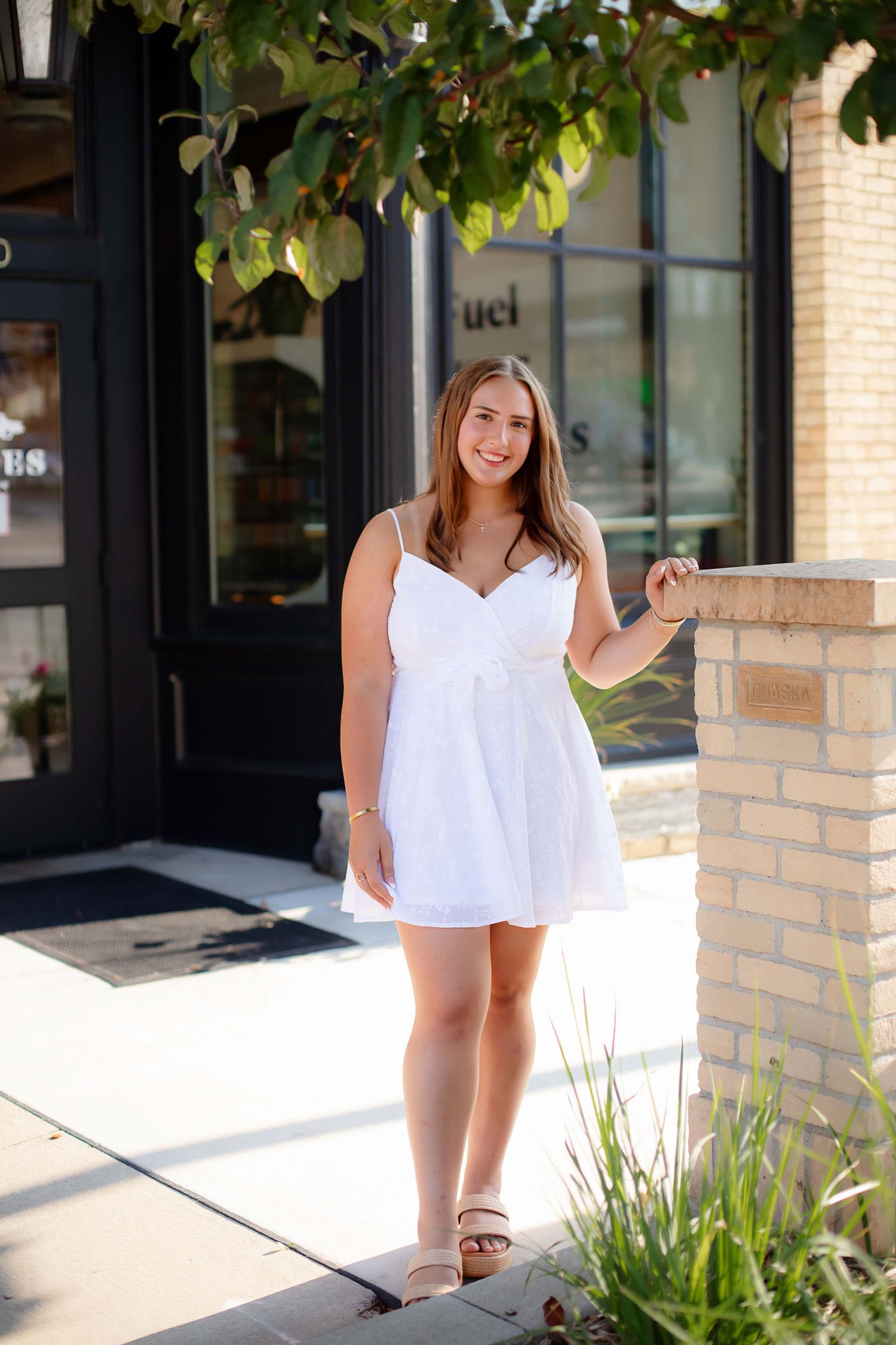 A person in a white sleeveless top poses in front of a brick building with greenery and windows.