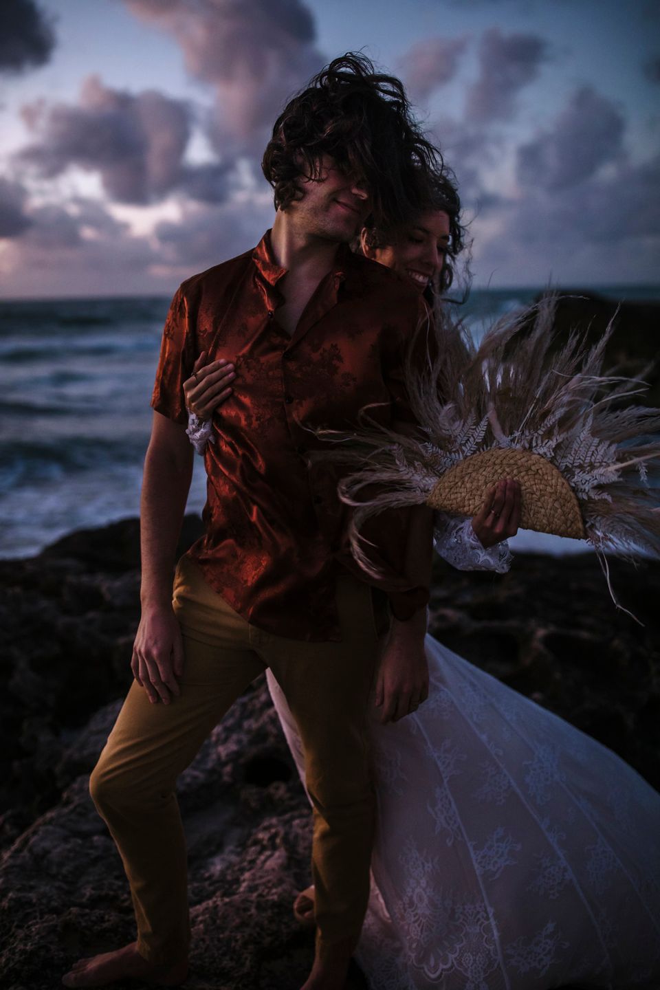 A couple in wedding attire poses playfully on coastal rocks during sunset with dramatic ocean waves.