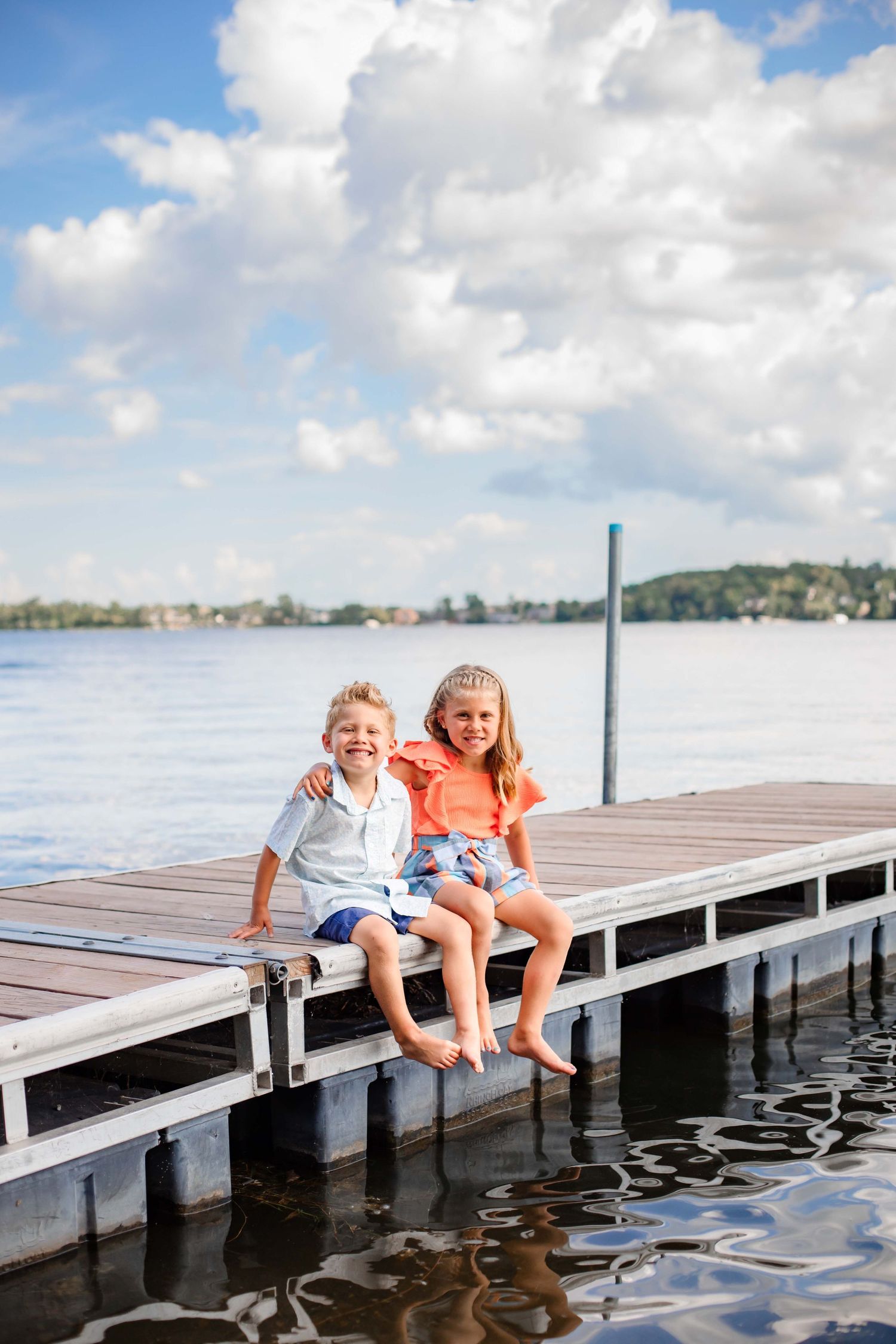 Two children sitting casually on wooden dock edge over lake waters.