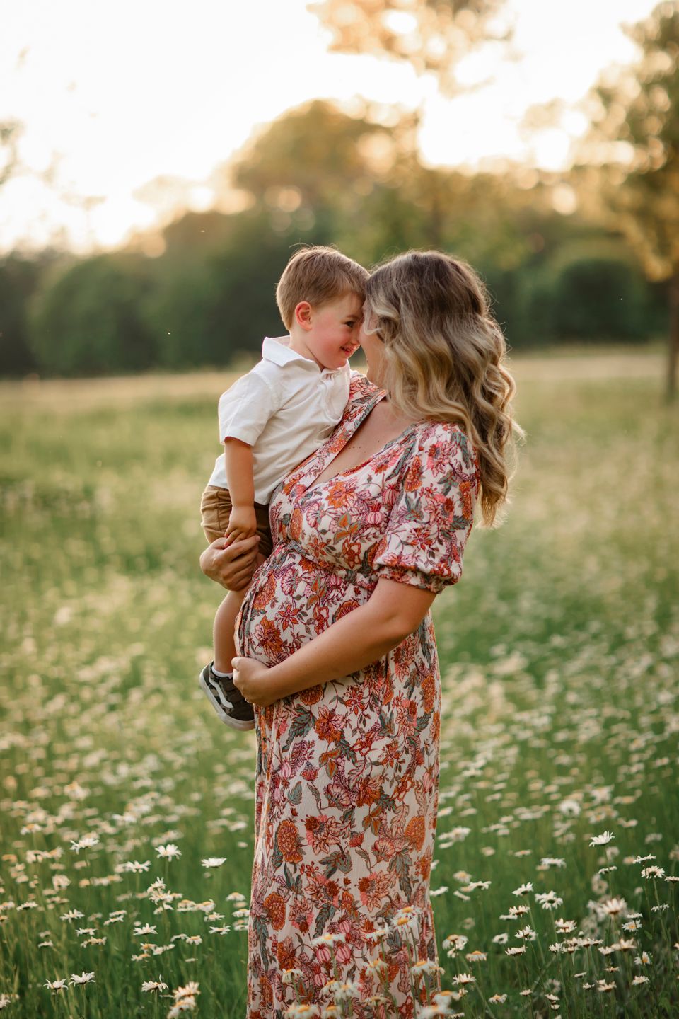 A tender moment captured between a woman in floral dress and toddler amid a field of wildflowers at sunset.