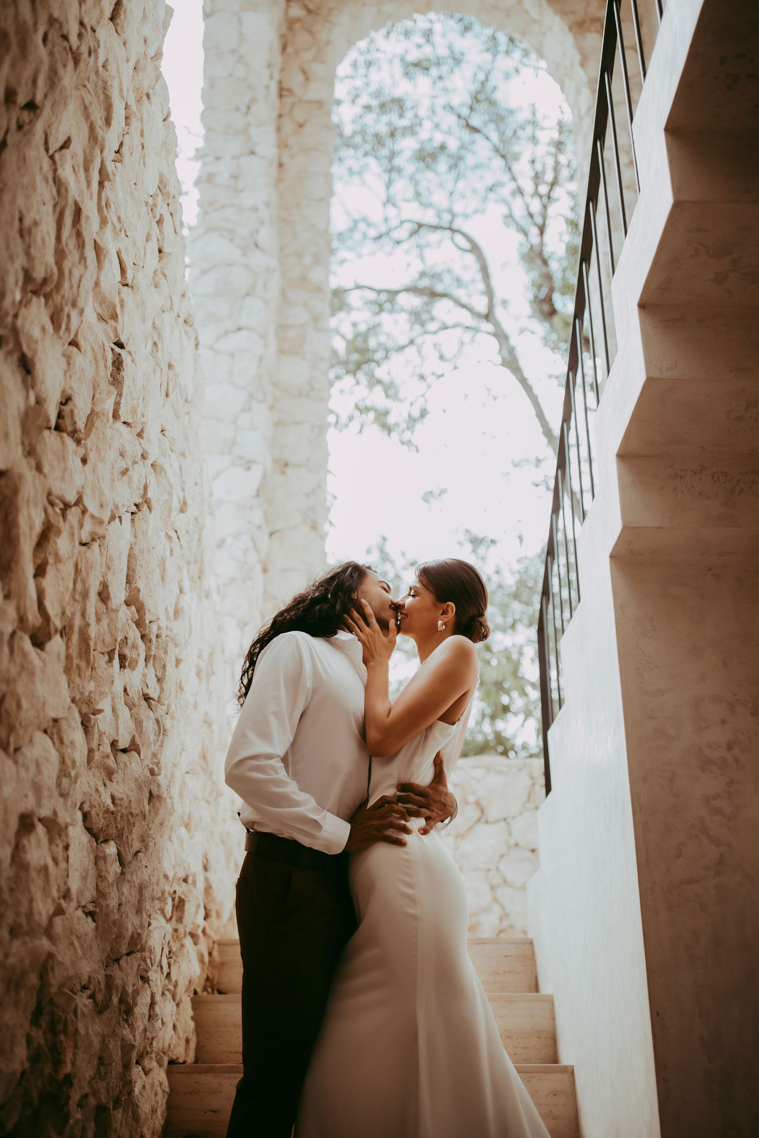 Romantic couple sharing a kiss on stone staircase with natural light streaming through.