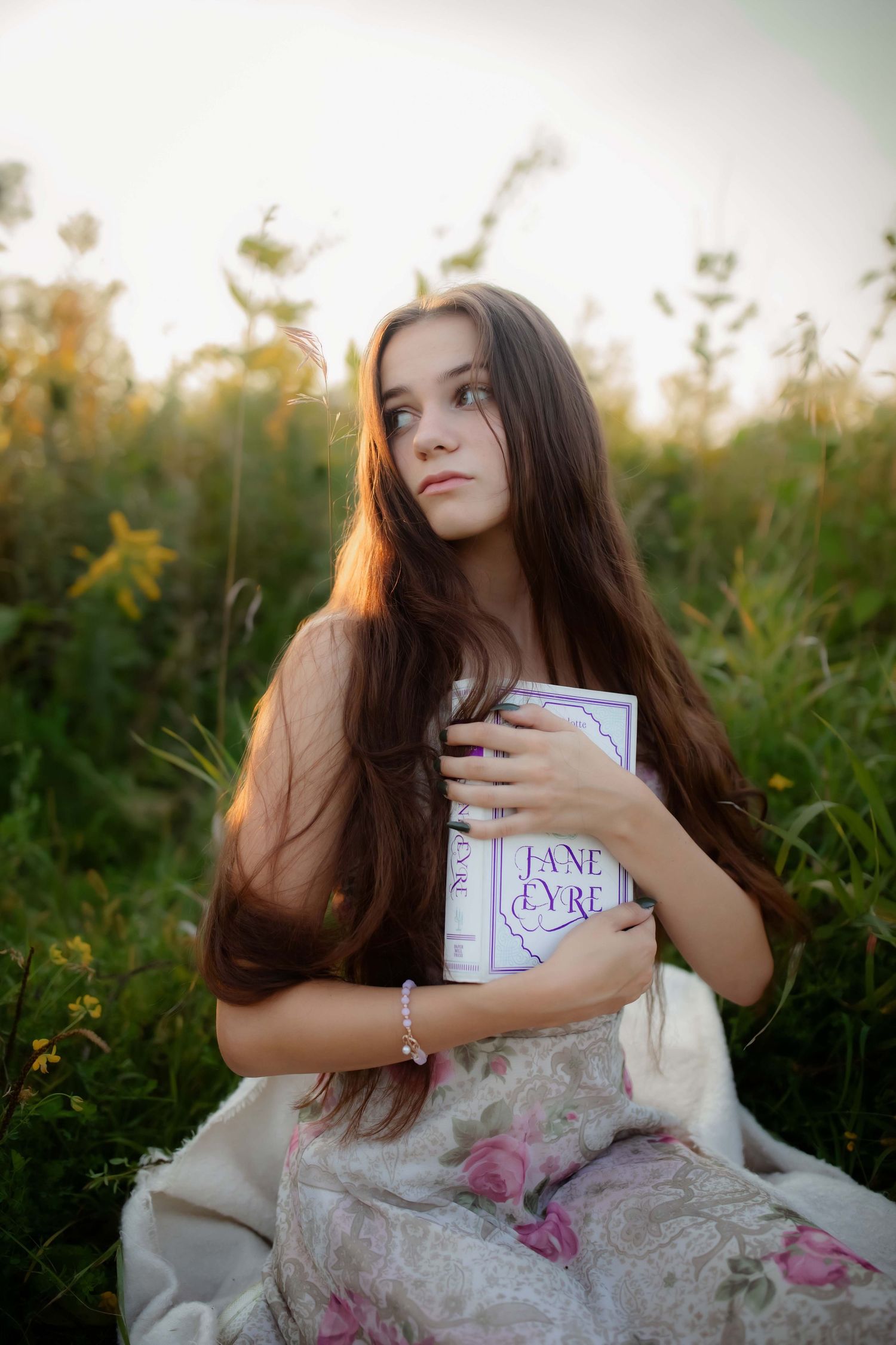 Dreamy outdoor portrait holding Jane Eyre book while wearing floral dress in golden meadow.