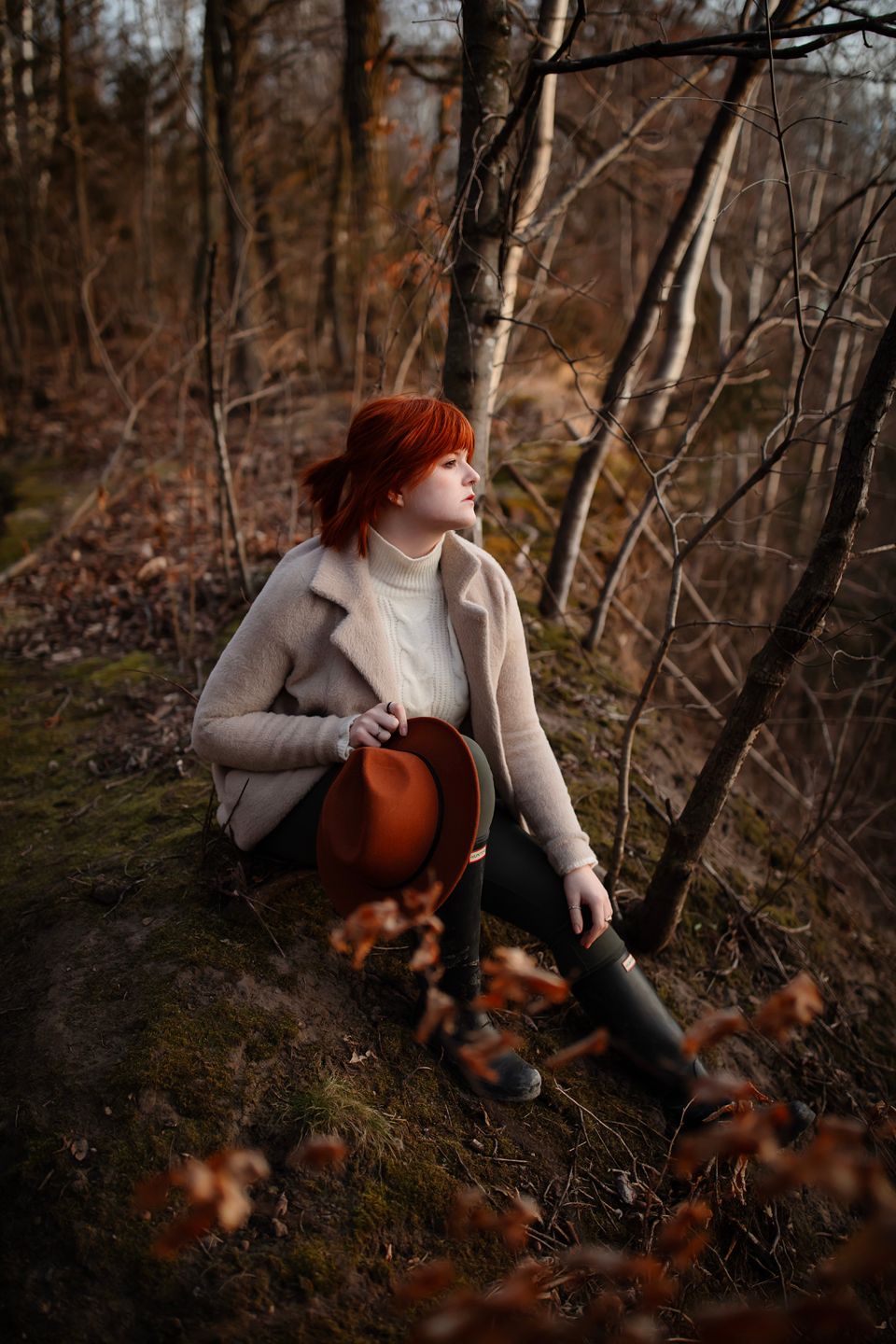 A figure in a beige coat and boots sits among autumn foliage in a forest setting.