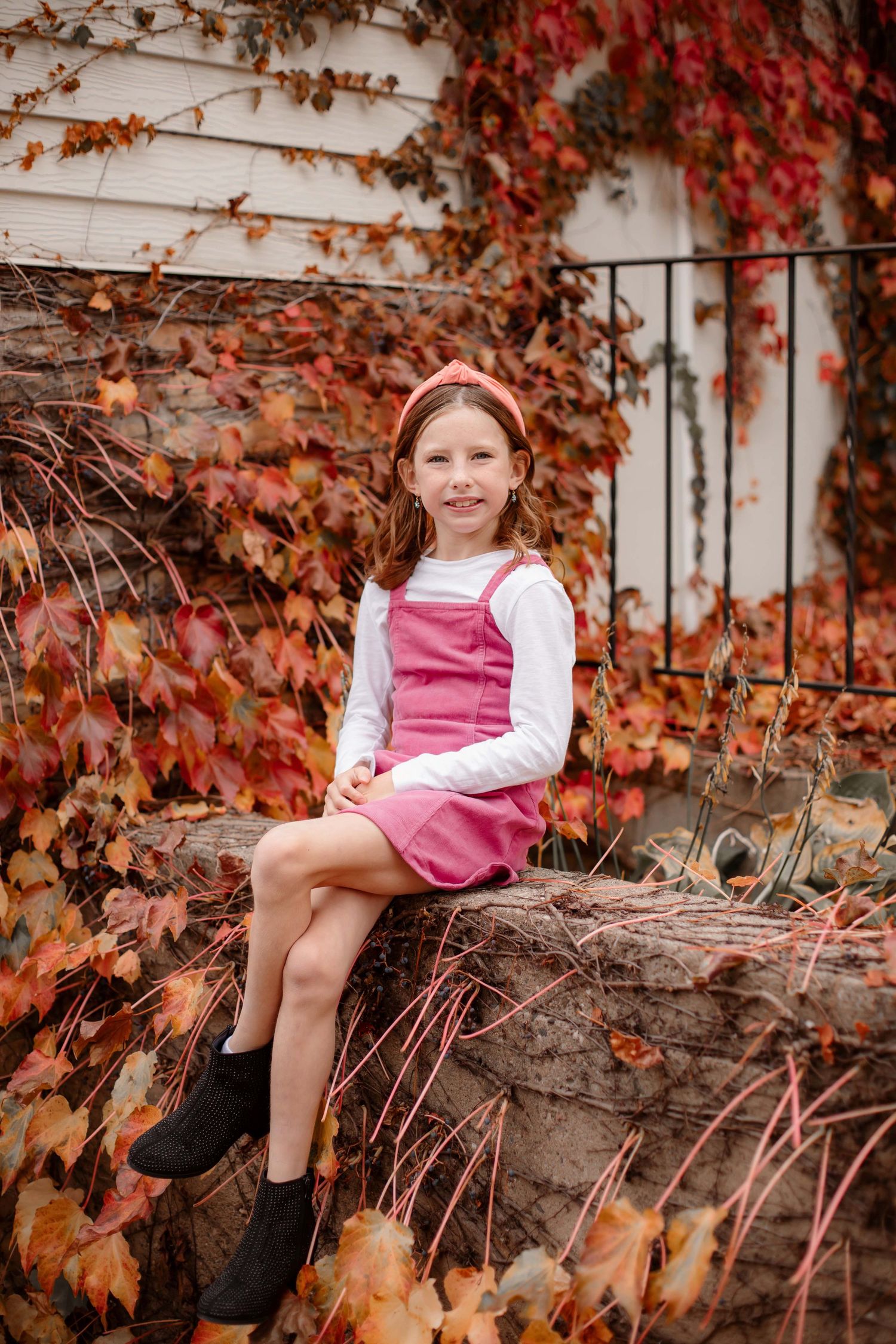 Autumn portrait with red ivy vines growing on white wall featuring pink pinafore dress and black boots.