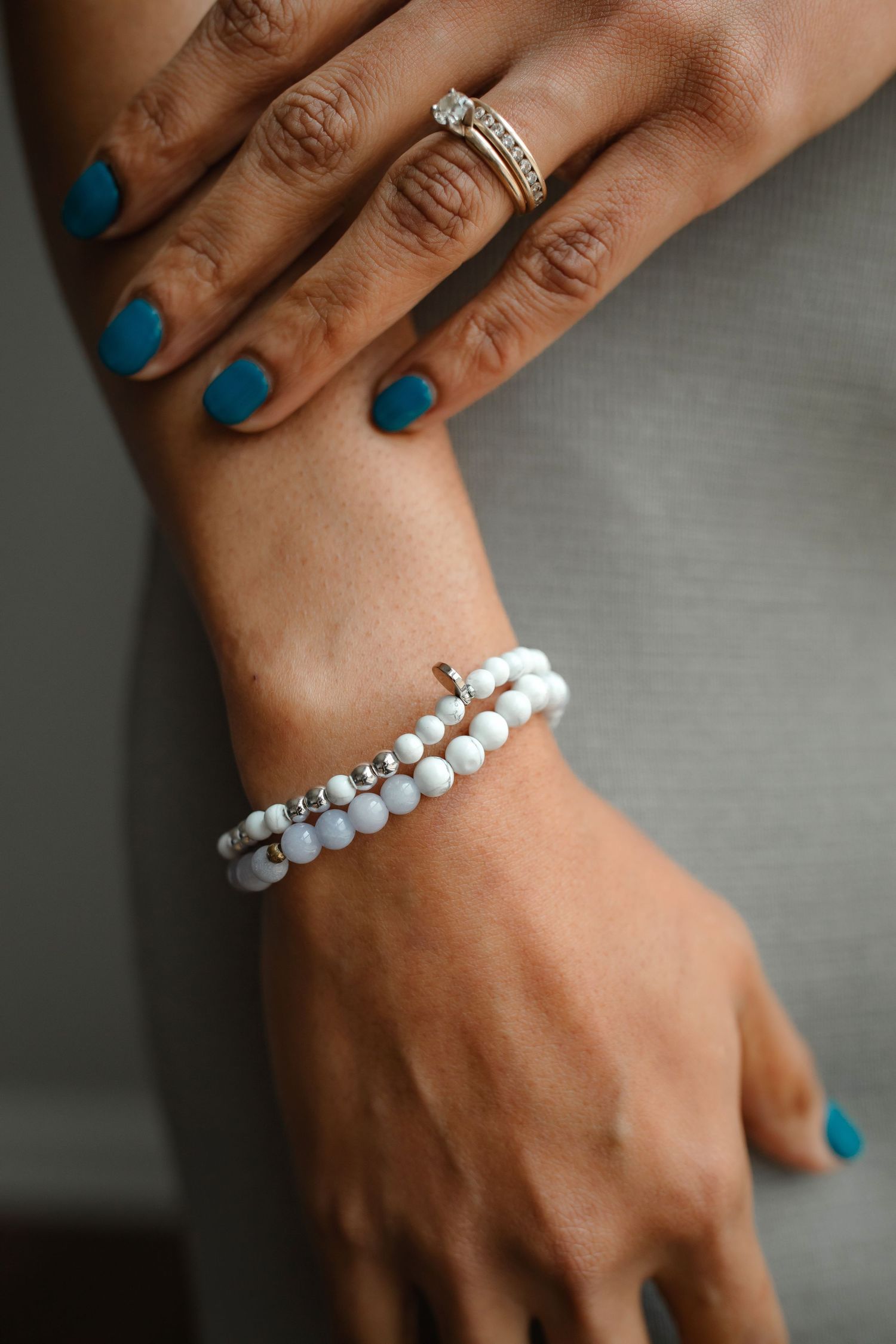 Hand with blue nails wearing diamond rings and white beaded bracelets.