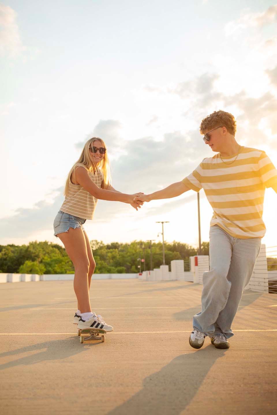 A young couple holds hands on a rooftop parking lot at sunset, with the girl balancing on a skateboard wearing denim shorts.