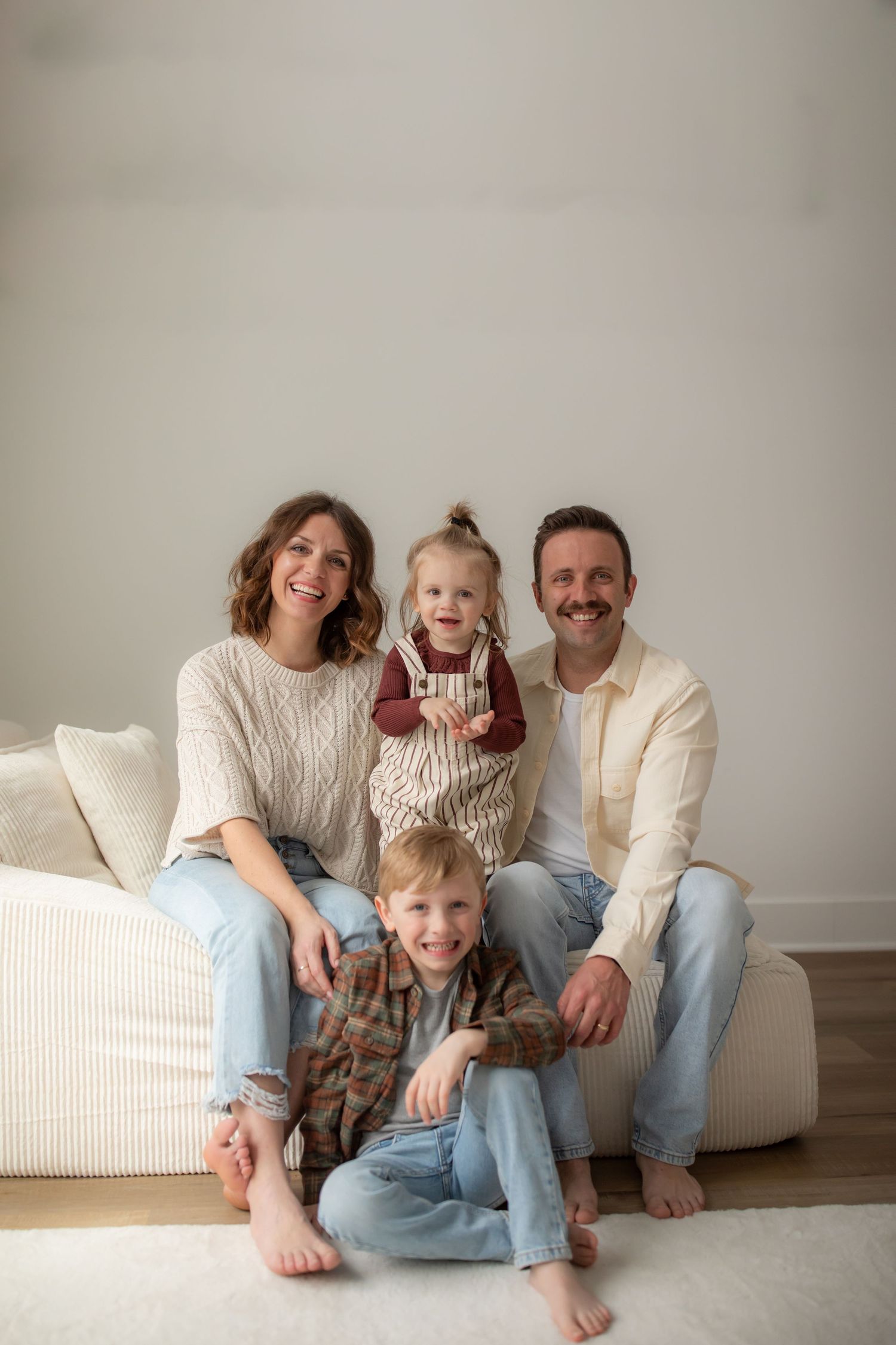 Family wearing light colored clothing sits together on white couch in casual lifestyle photoshoot.
