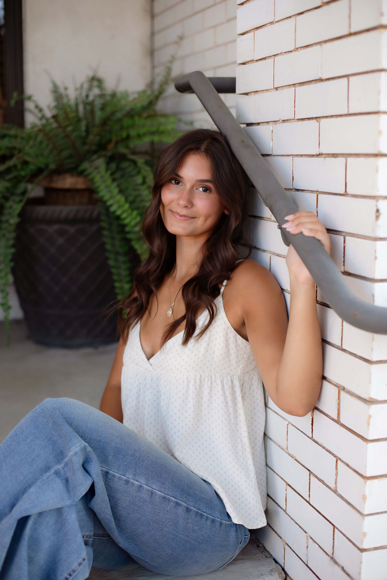 Young woman in white tank top and jeans sitting against a white brick wall, smiling warmly at the camera.