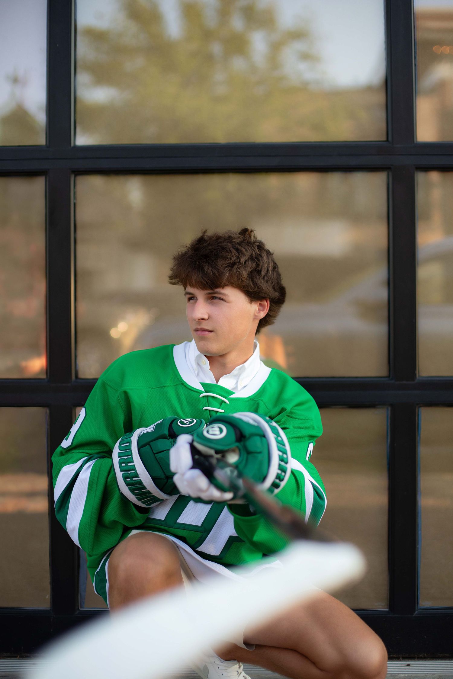Young hockey player in green jersey holding stick and gloves, seated by large window in senior portrait photo.