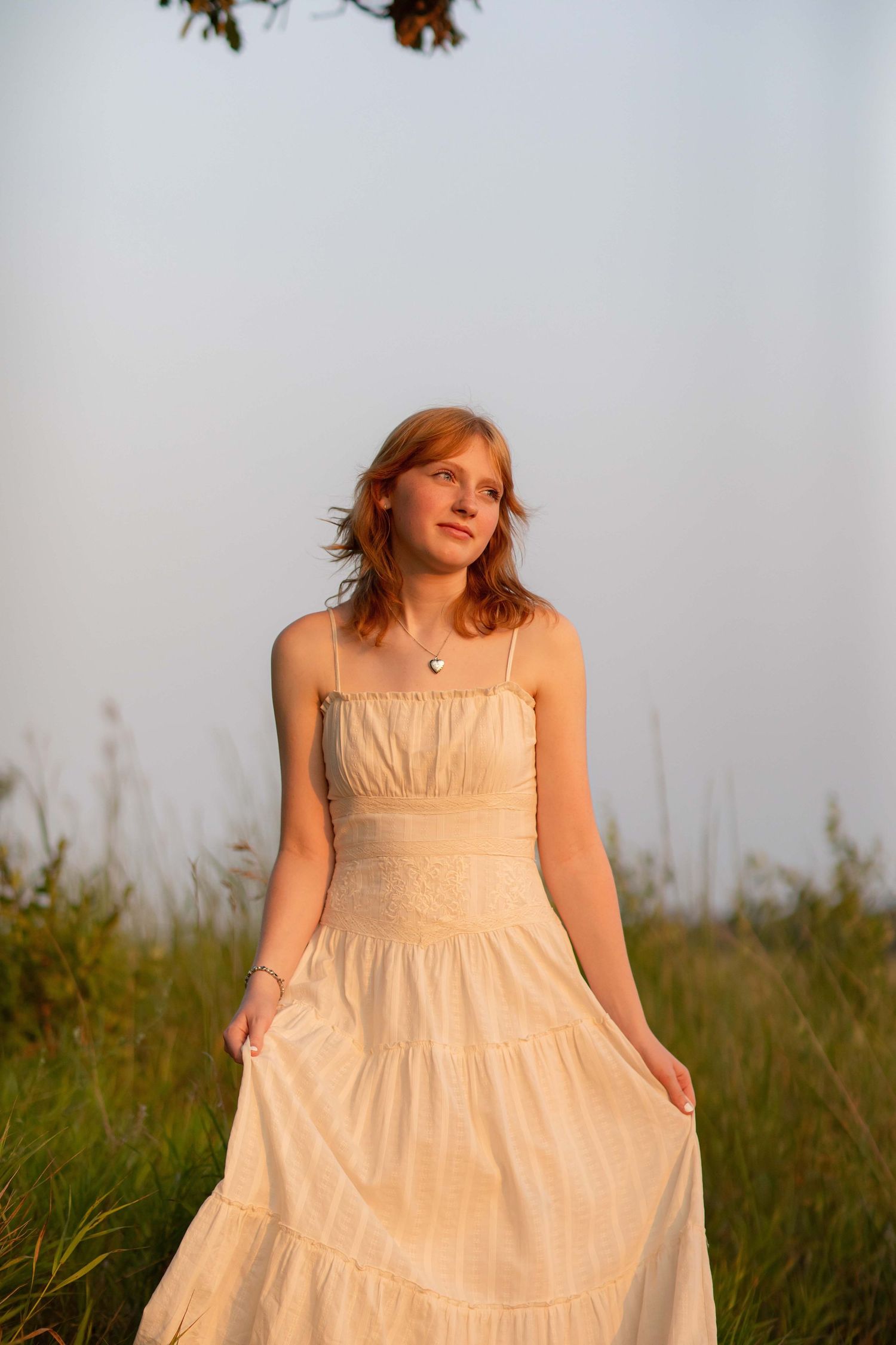 Young woman in cream sundress standing in green field during golden hour sunset light.