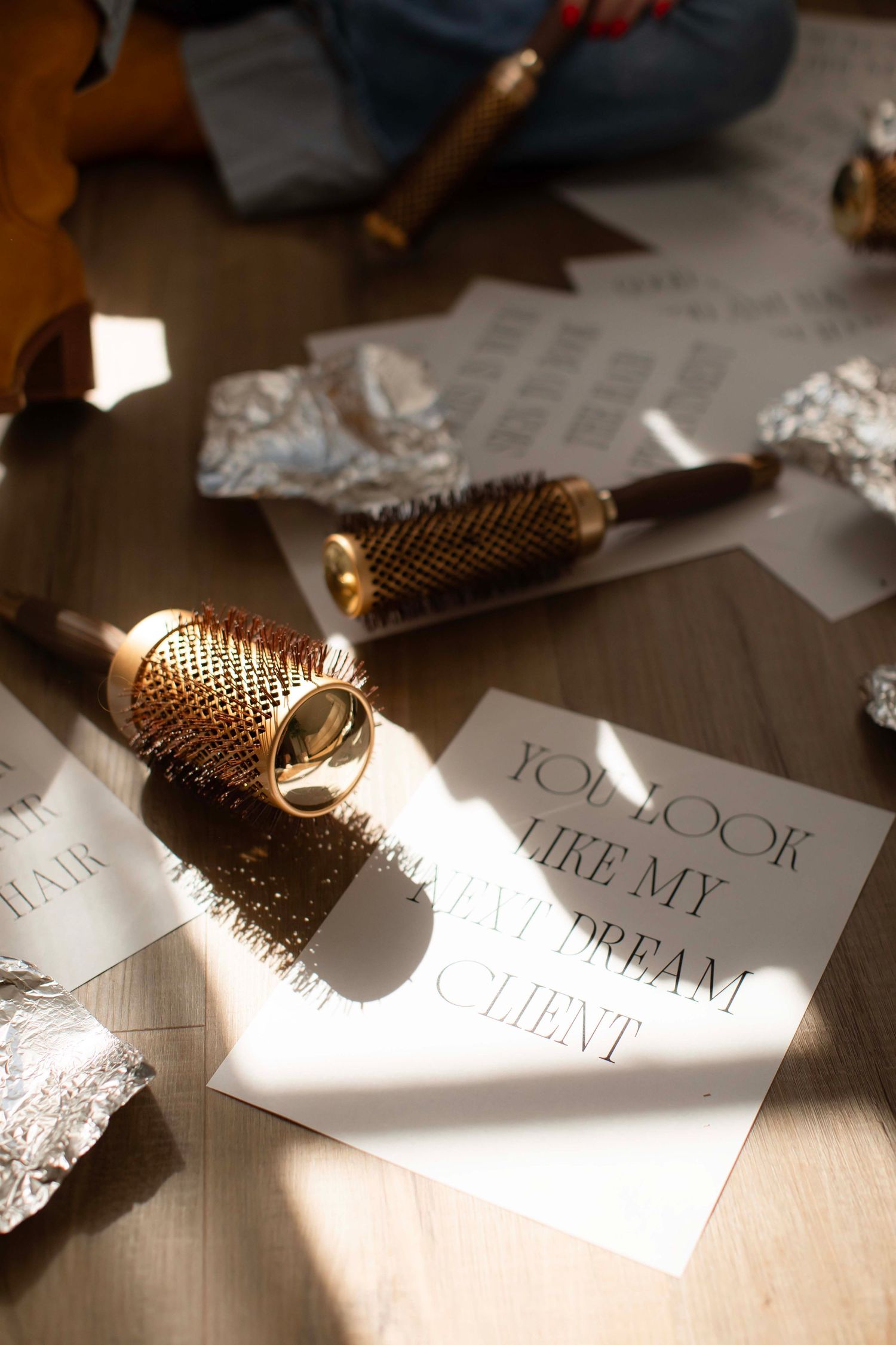 Gold round brushes and foil highlights on wooden surface with hairstylist motivational cards in warm sunlight.