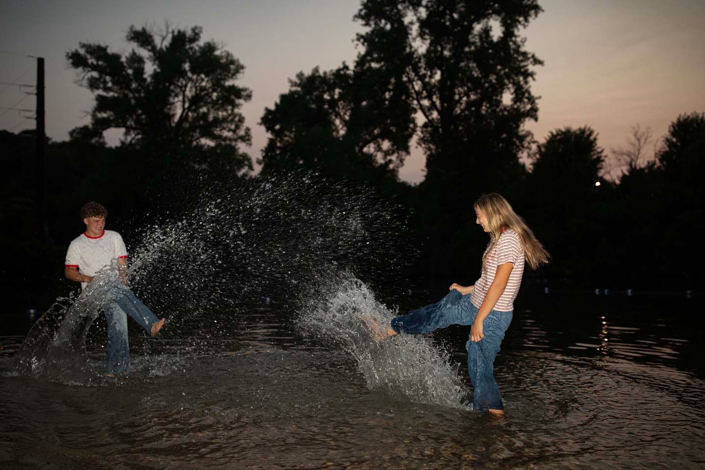 Two people splashing in shallow water at dusk, wearing jeans, enjoying a playful moment near tree-lined shore.