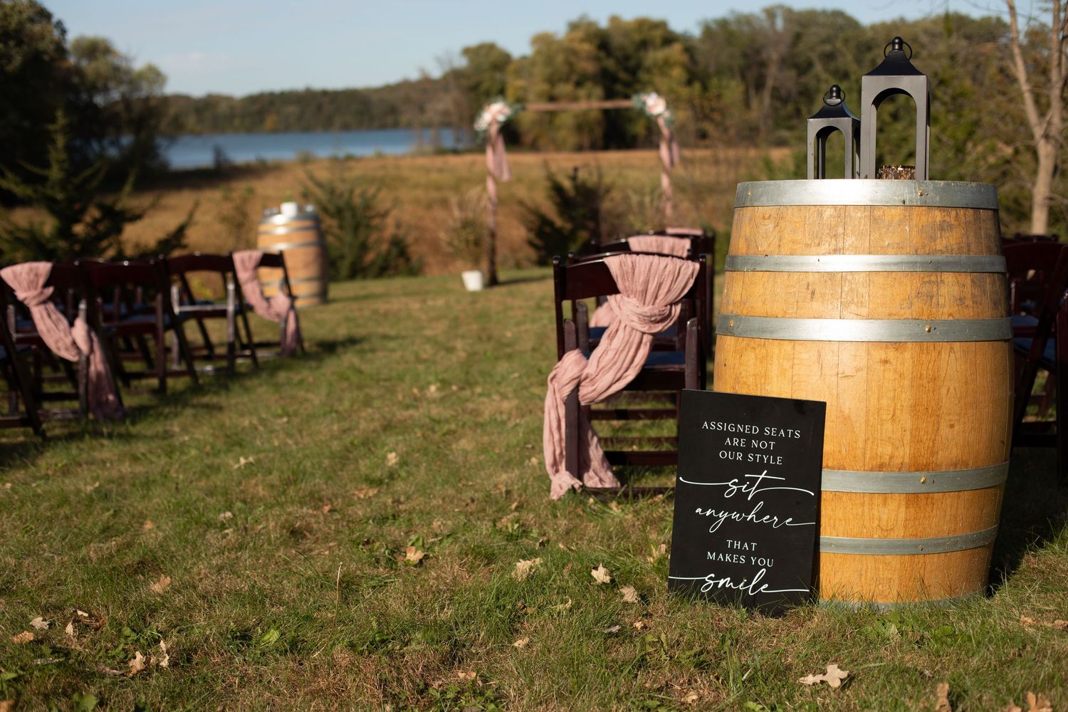 Rustic wedding decor with wooden barrels and pink chairs arranged outdoors near a lake.