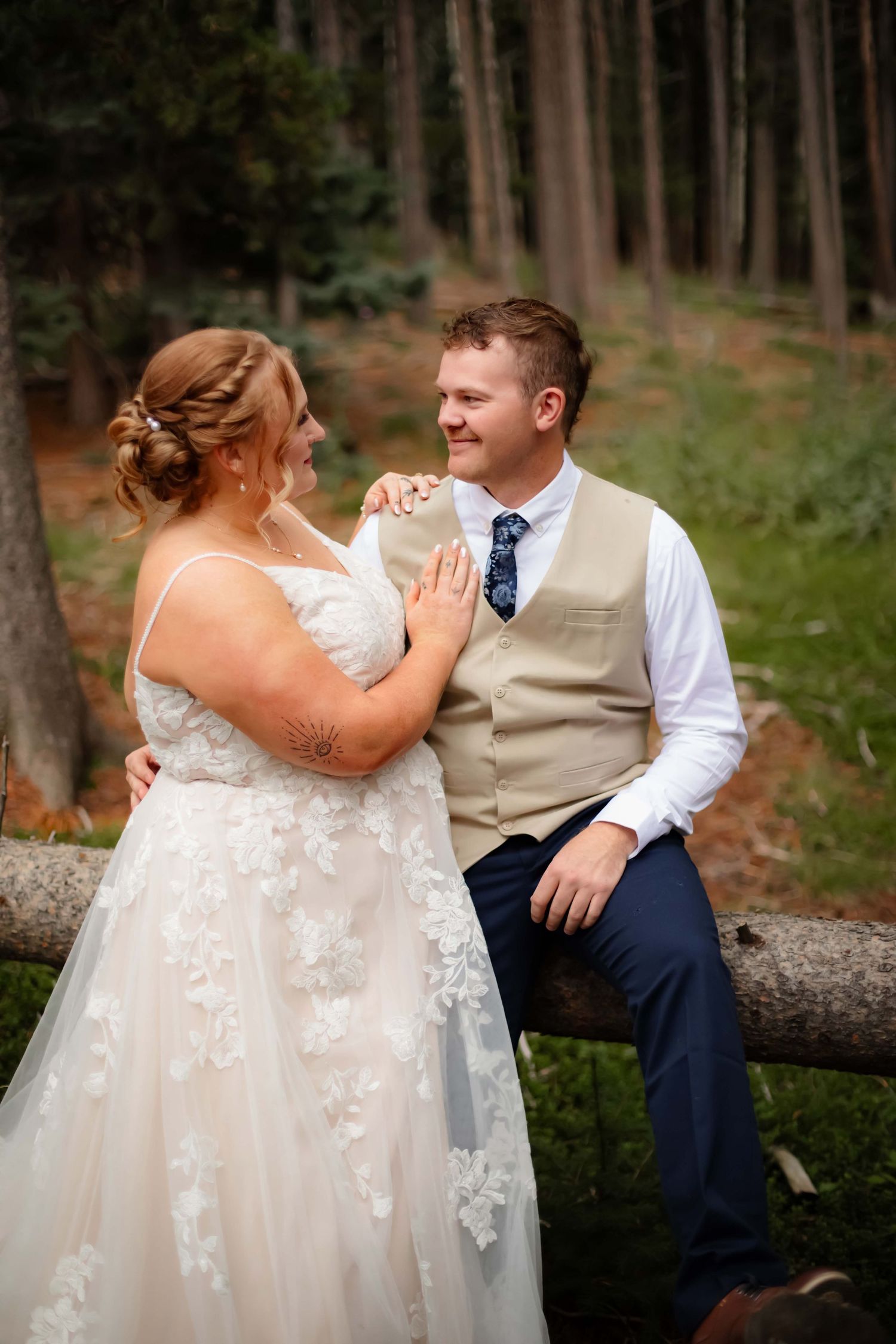 A romantic moment between newlyweds sitting on a fallen log in a forest setting during their wedding photoshoot.