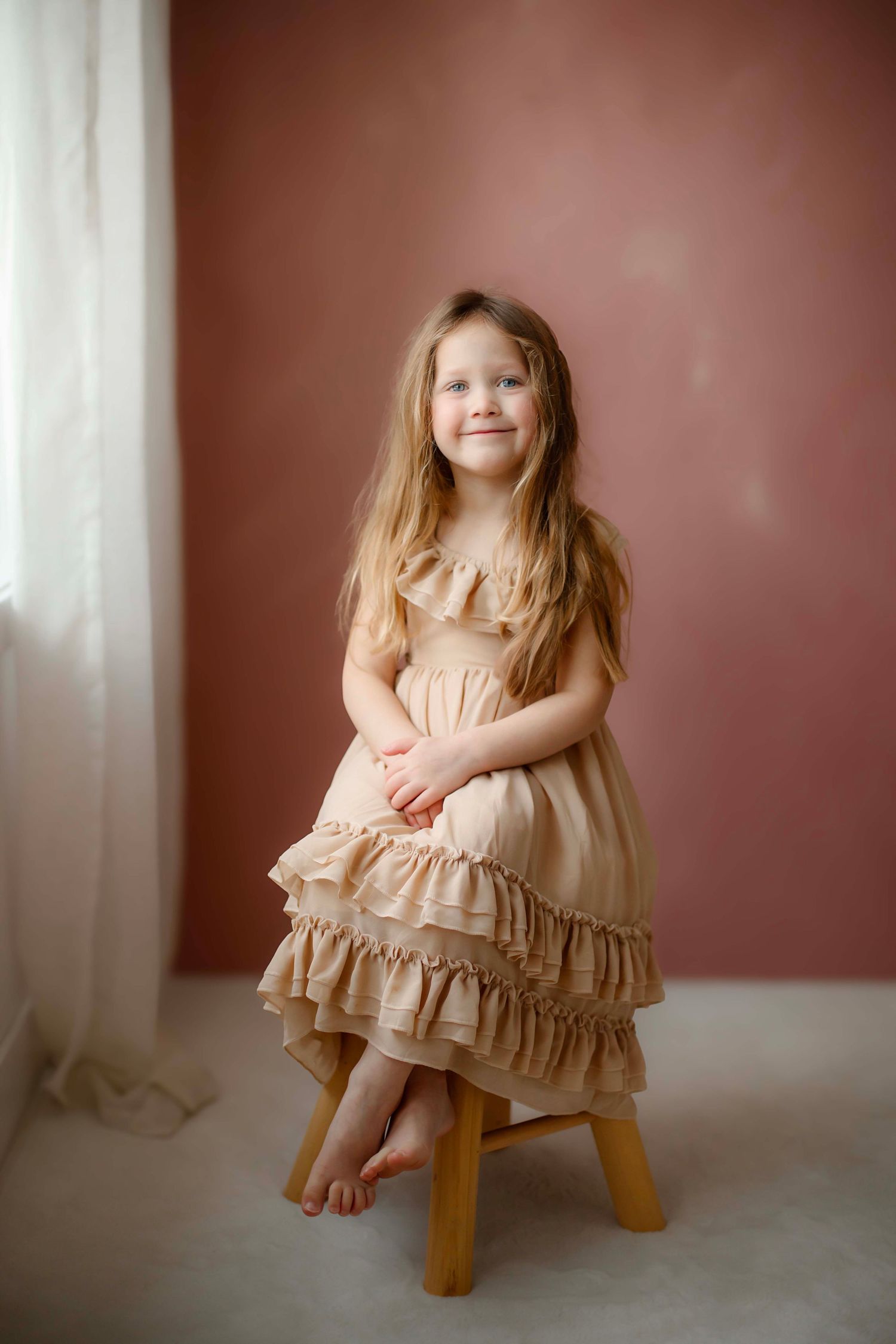A child in a peach ruffled dress sits on a wooden stool against a burgundy wall with soft window light.