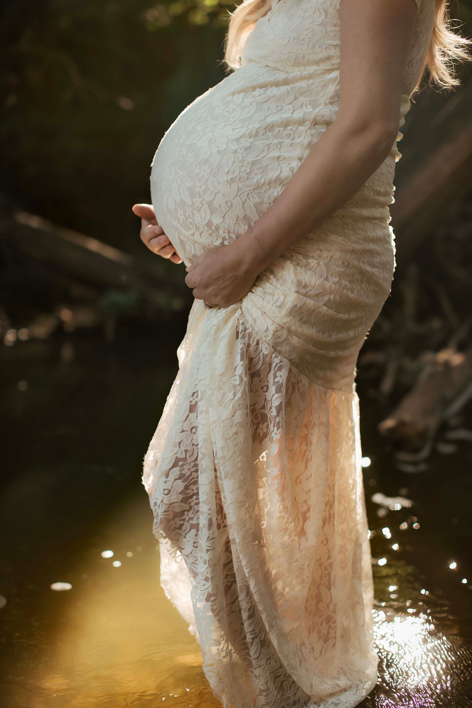Pregnant woman in white lace dress cradling baby bump outdoors near water in golden sunlight.