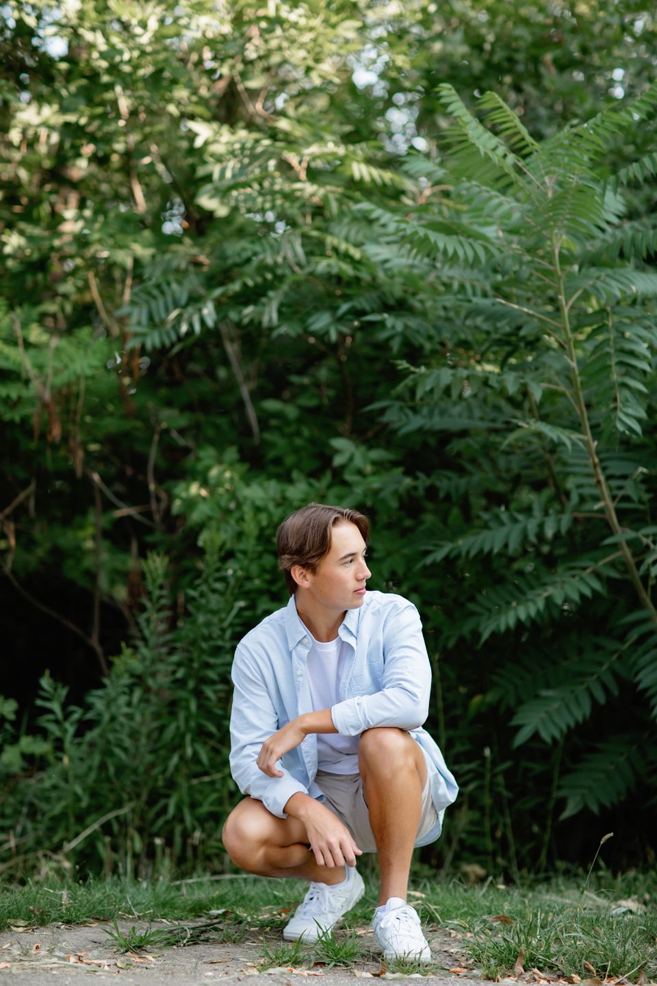 Figure in light blue shirt and shorts sits crouched in a garden setting with lush summertime foliage in background.