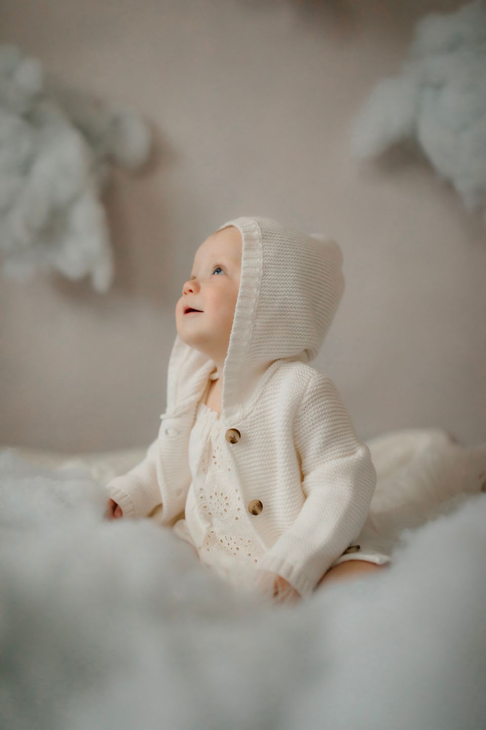 Baby wearing cream hooded sweater sitting among cloud-like decorations.