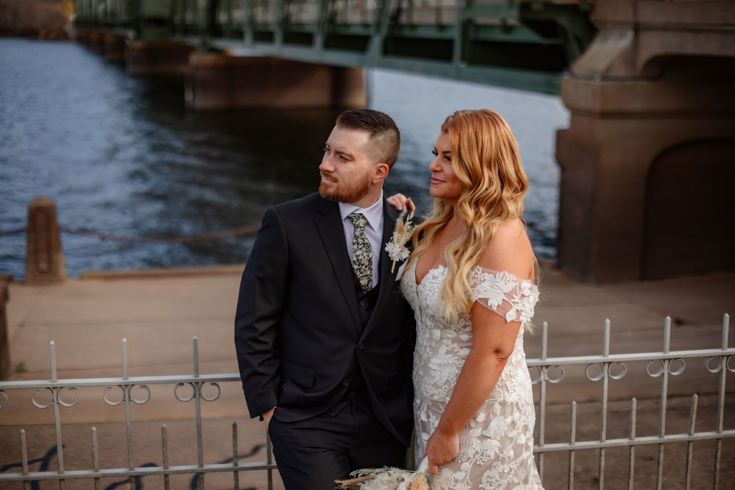 A wedding couple stands by a waterfront with an industrial bridge in the background.