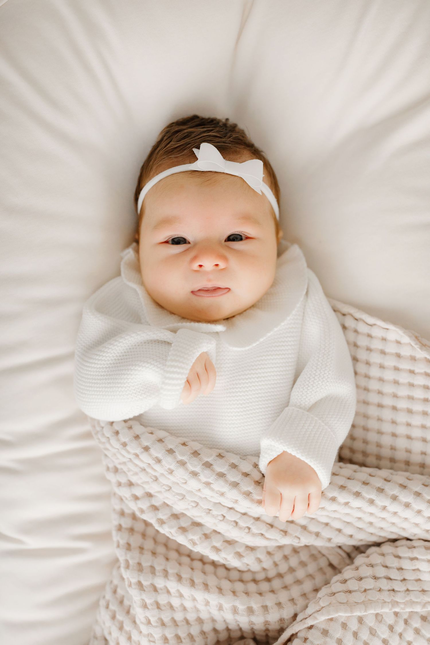 Newborn baby wearing white sweater and headband lying on soft white bedding.