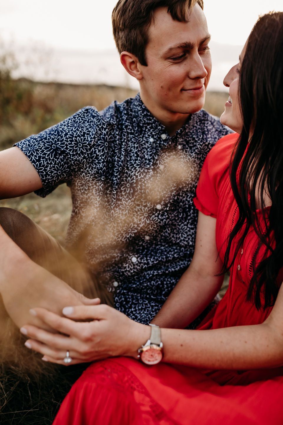 Intimate moment between couple wearing blue patterned shirt and red dress in natural setting.