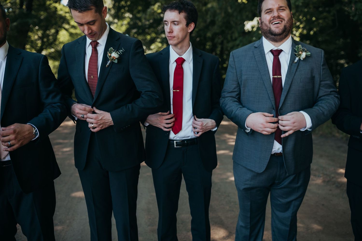 Groomsmen in dark suits with red ties standing together outdoors.