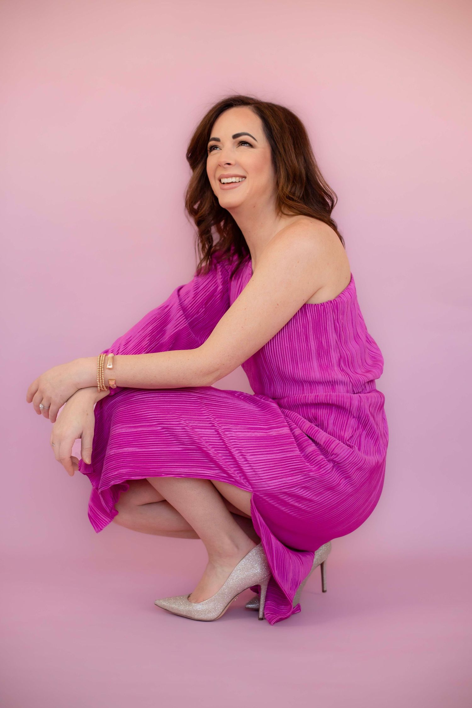 Crouching pose in magenta pleated dress with silver heels against pink background.