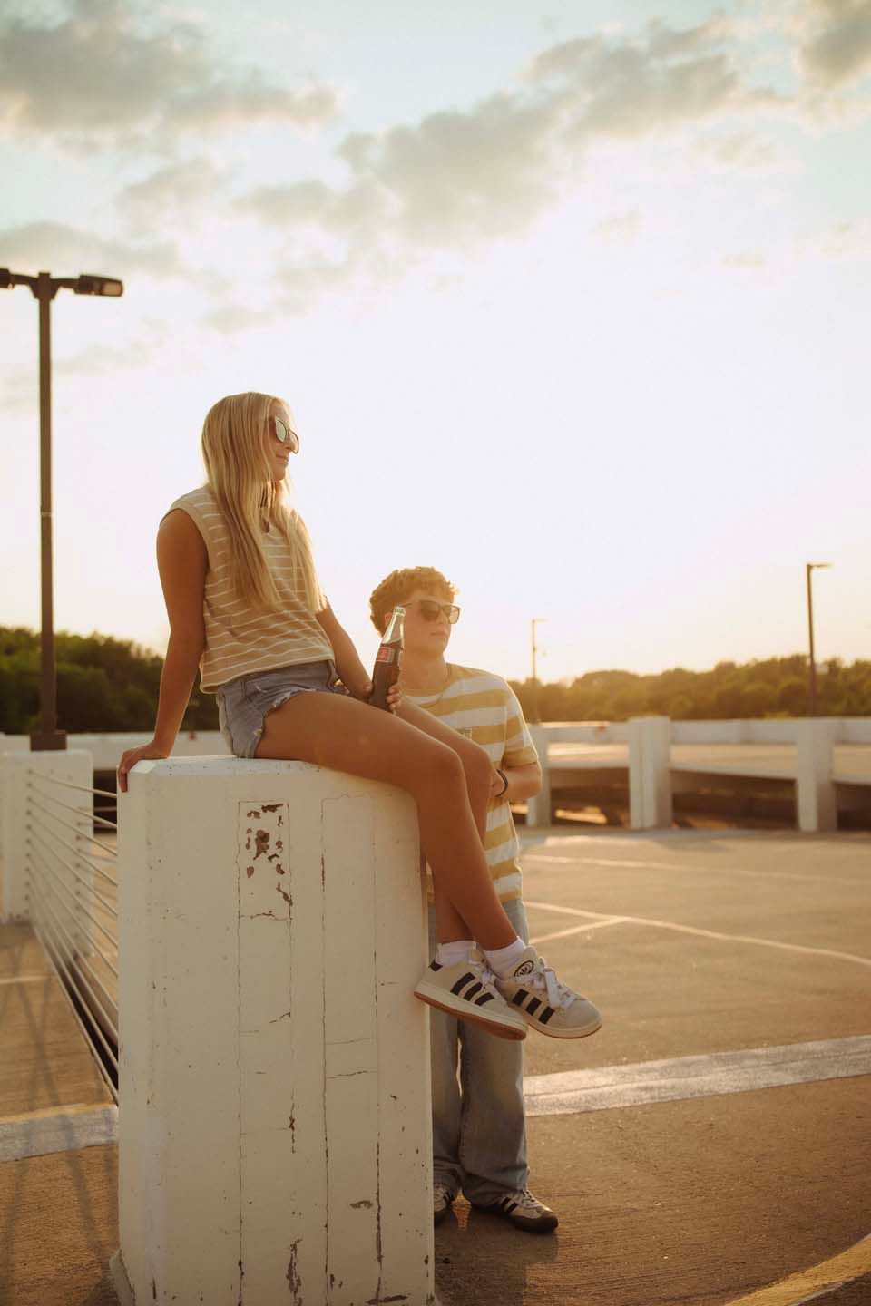 Two young friends sit and stand on a white concrete pillar in a sunny parking lot during golden hour sunset.