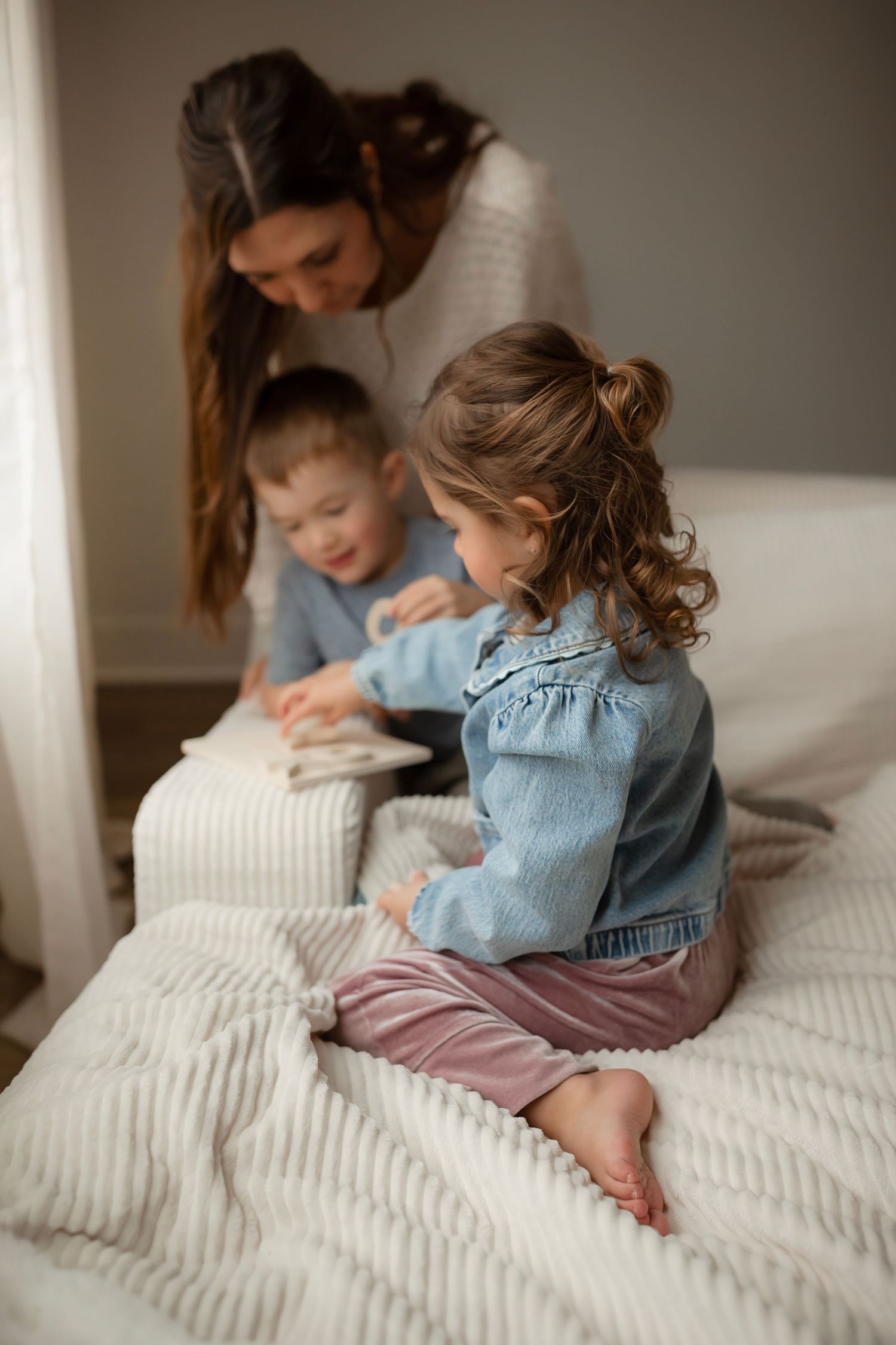Intimate family scene with children in denim clothing sitting together on white bedding in natural light.