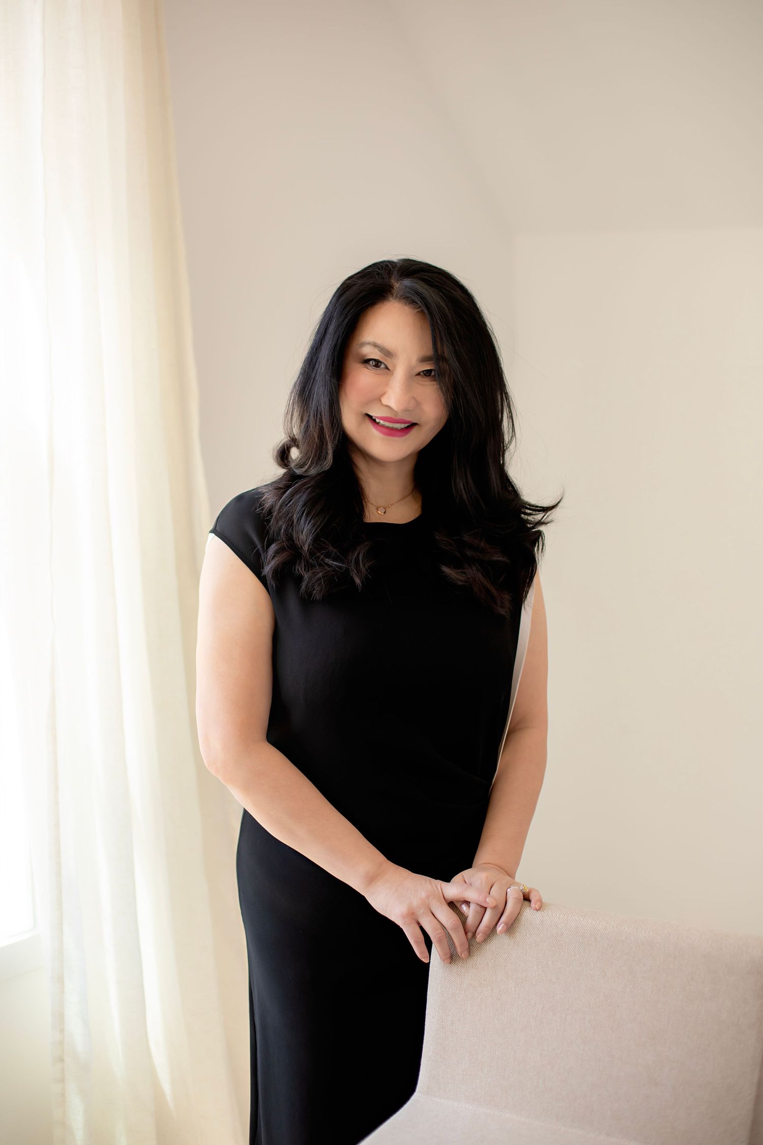 A person in a black dress poses confidently against a light-colored wall in a minimalist setting.